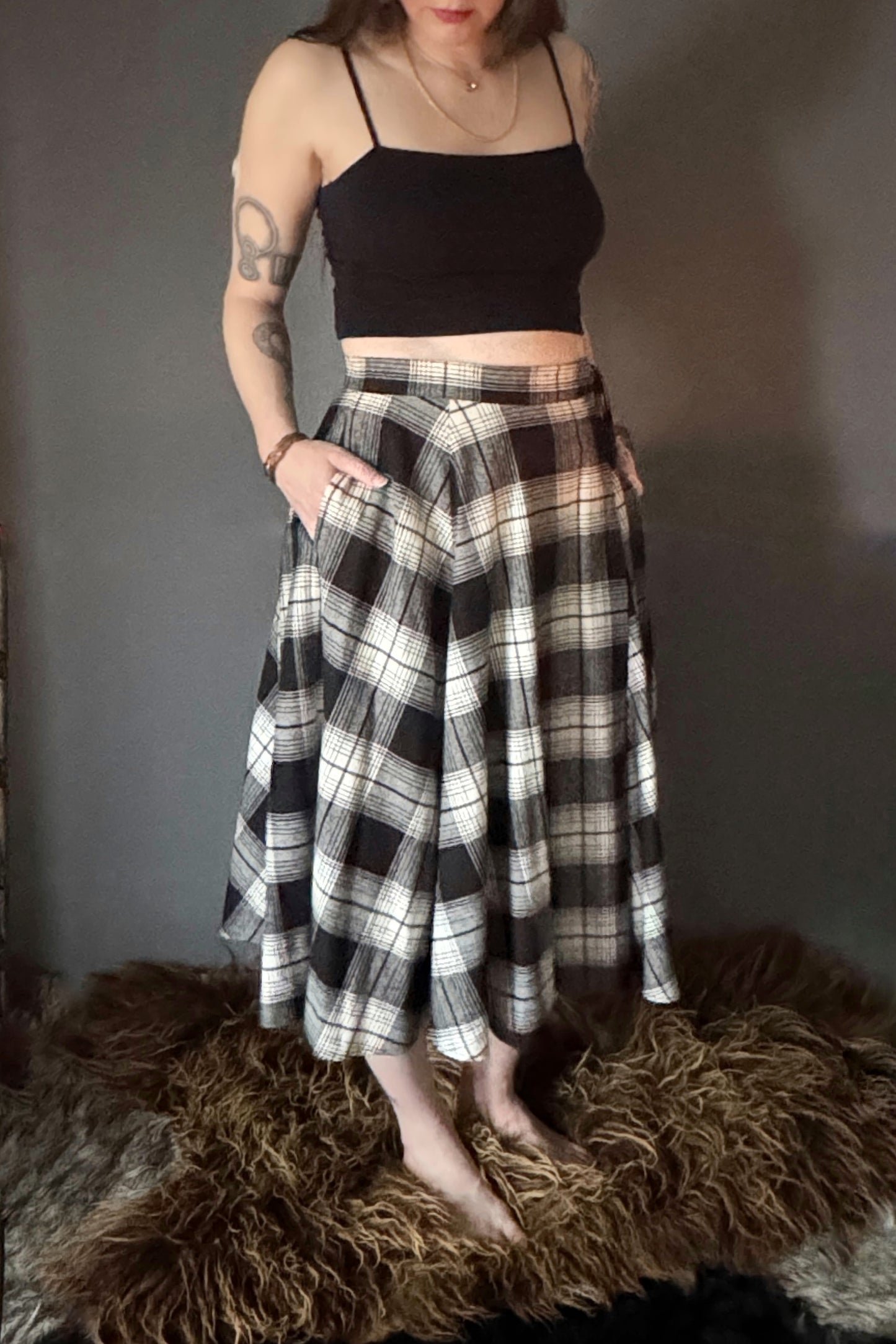 A photo of a black and white plaid wrap skirt. Only the lower half the body is showing featuring the mid calf length, side pockets and long ties. An adjustable cotton tartan wrap skirt, size inclusive and made in Canada.