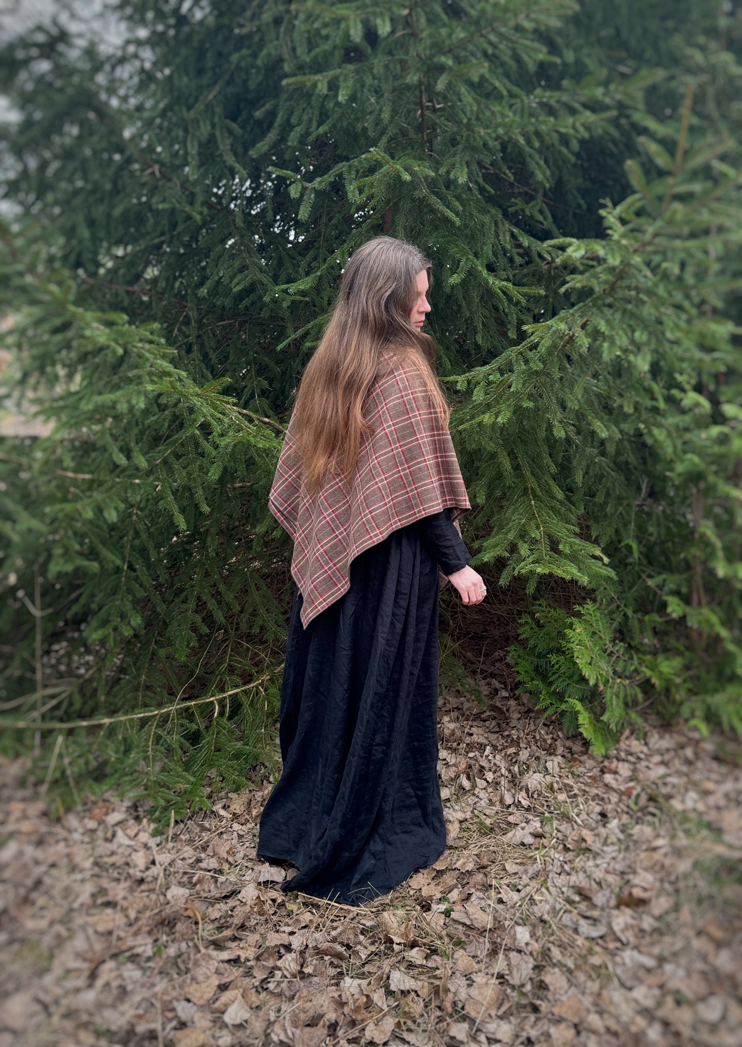 A celtic woman stands in front some evergreen trees wearing a black tunic dress with a brown long belt and featuring an earth toned plaid shawl. Brown undertones with white and red stripes. in this photo it is a back side view of the shawl draped over the shoulders and closed at the front. You can see the point of the shawl and size. Historical costume, ancestor reverence, old gods, made in canada.