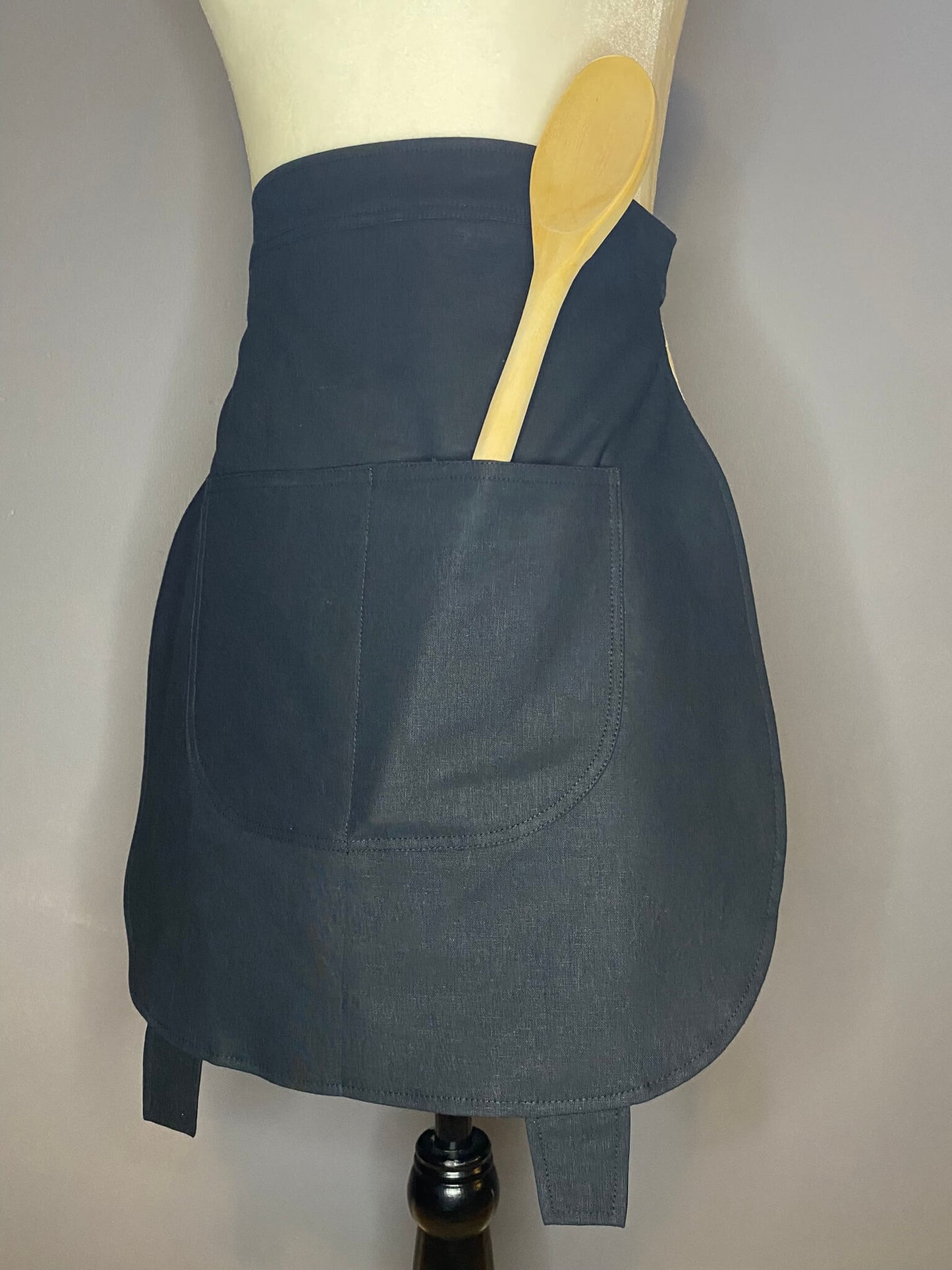 A black cotton linen apron shaped like a cauldron. Featured here with a wooden spoon in one of the front pockets.