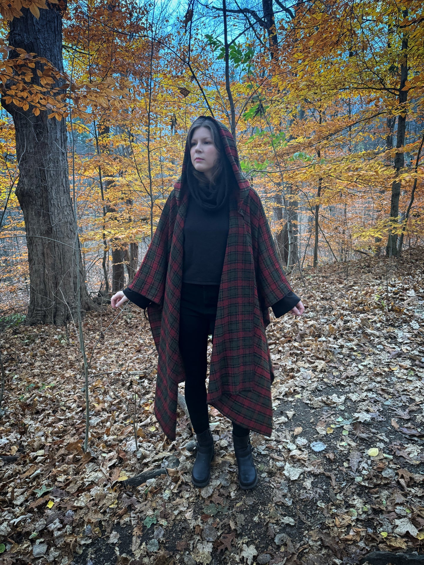 A witch standing in a fprest covered in fallen leaves wearing black pants and a black hexen hoodie with a tartan poncho layered over top. The hoodie features a large cowl hood and cropped just at top of the hips. Featuring slight bell sleeves and its made from a soft bamboo knit with a fleece interior. Very soft and warm good for layering. Witch clothing, fall wear, sustainably made in canada