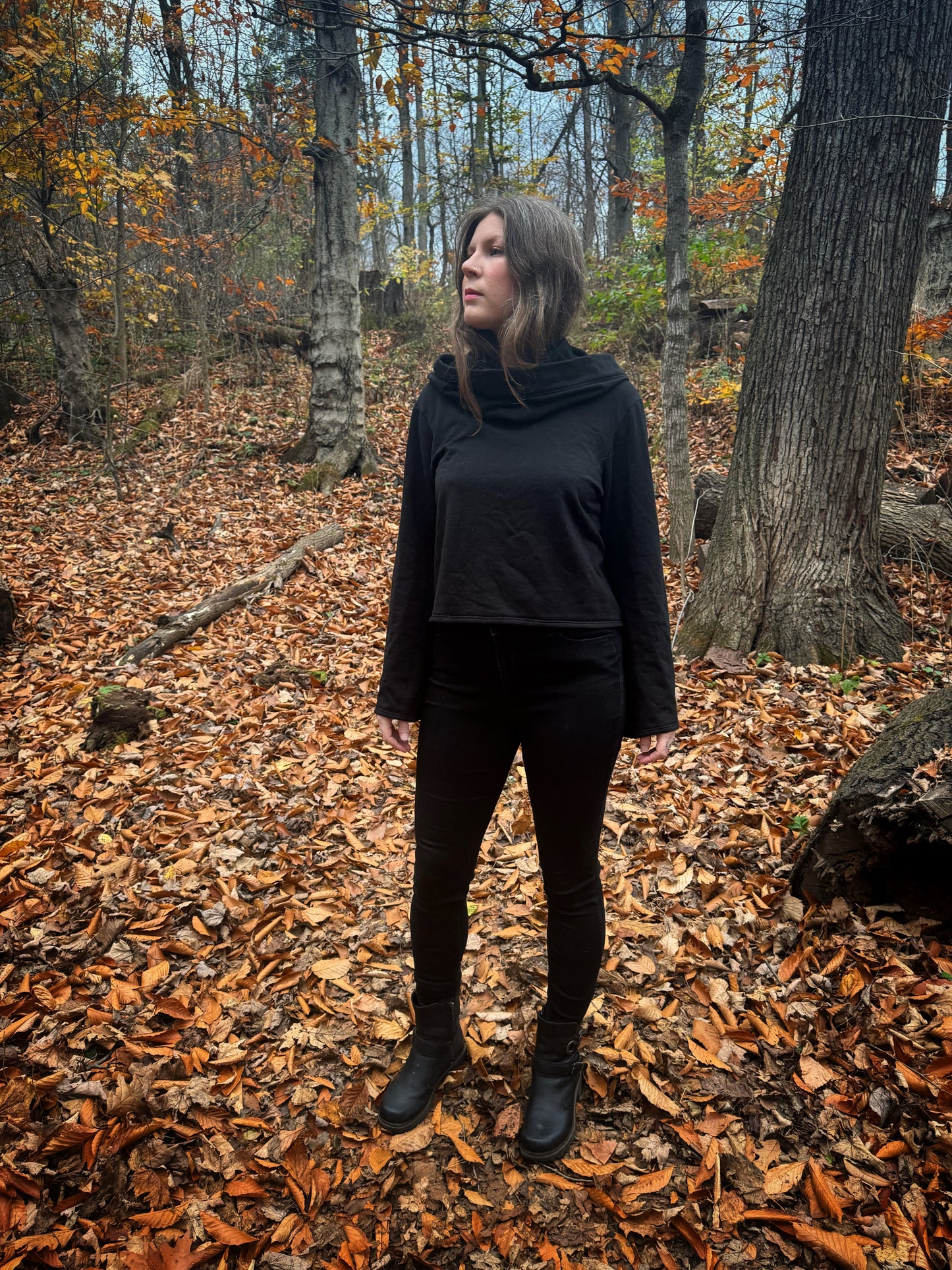 A witch standing in a forest covered in fallen leaves wearing black pants and a black hexen hoodie. The hoodie features a large cowl hood and cropped just at top of the hips. Featuring slight bell sleeves and its made from a soft bamboo knit with a fleece interior. Very soft and warm good for layering. Witch clothing, fall wear, sustainainably made in canada