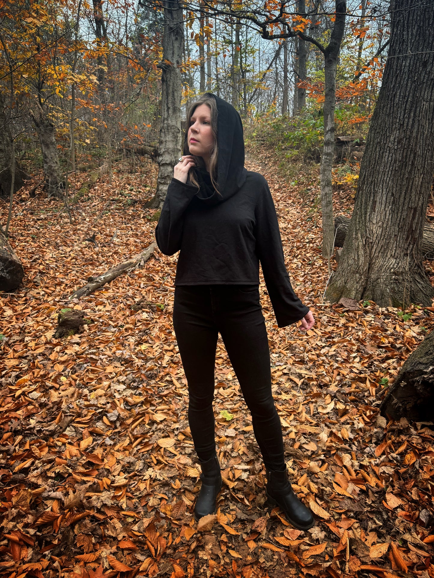 A witch standing in a fprest covered in fallen leaves wearing black pants and a black hexen hoodie. The hoodie features a large cowl hood and cropped just at top of the hips. Featuring slight bell sleeves and its made from a soft bamboo knit with a fleece interior. Very soft and warm good for layering. Witch clothing, fall wear, sustainably made in canada