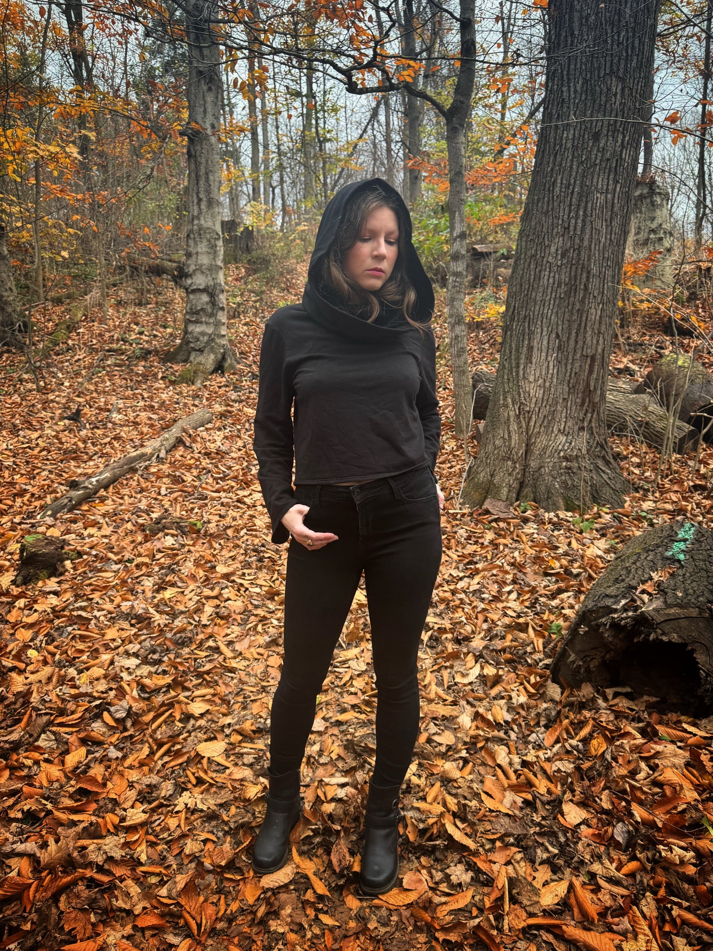 A witch standing in a fprest covered in fallen leaves wearing black pants and a black hexen hoodie. The hoodie features a large cowl hood and cropped just at top of the hips. Featuring slight bell sleeves and its made from a soft bamboo knit with a fleece interior. Very soft and warm good for layering. Witch clothing, fall wear, sustainably made in canada