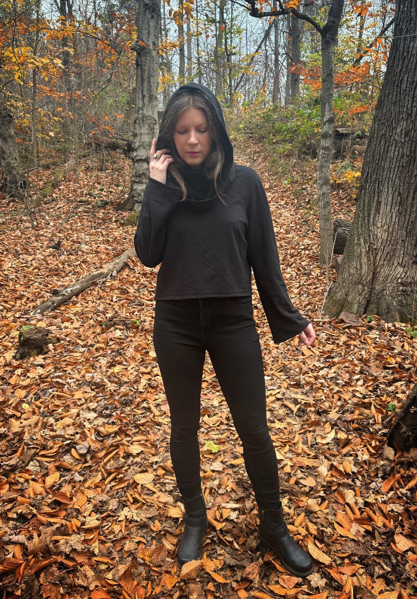 A witch standing in a fprest covered in fallen leaves wearing black pants and a black hexen hoodie. The hoodie features a large cowl hood and cropped just at top of the hips. Featuring slight bell sleeves and its made from a soft bamboo knit with a fleece interior. Very soft and warm good for layering. Witch clothing, fall wear, sustainably made in canada