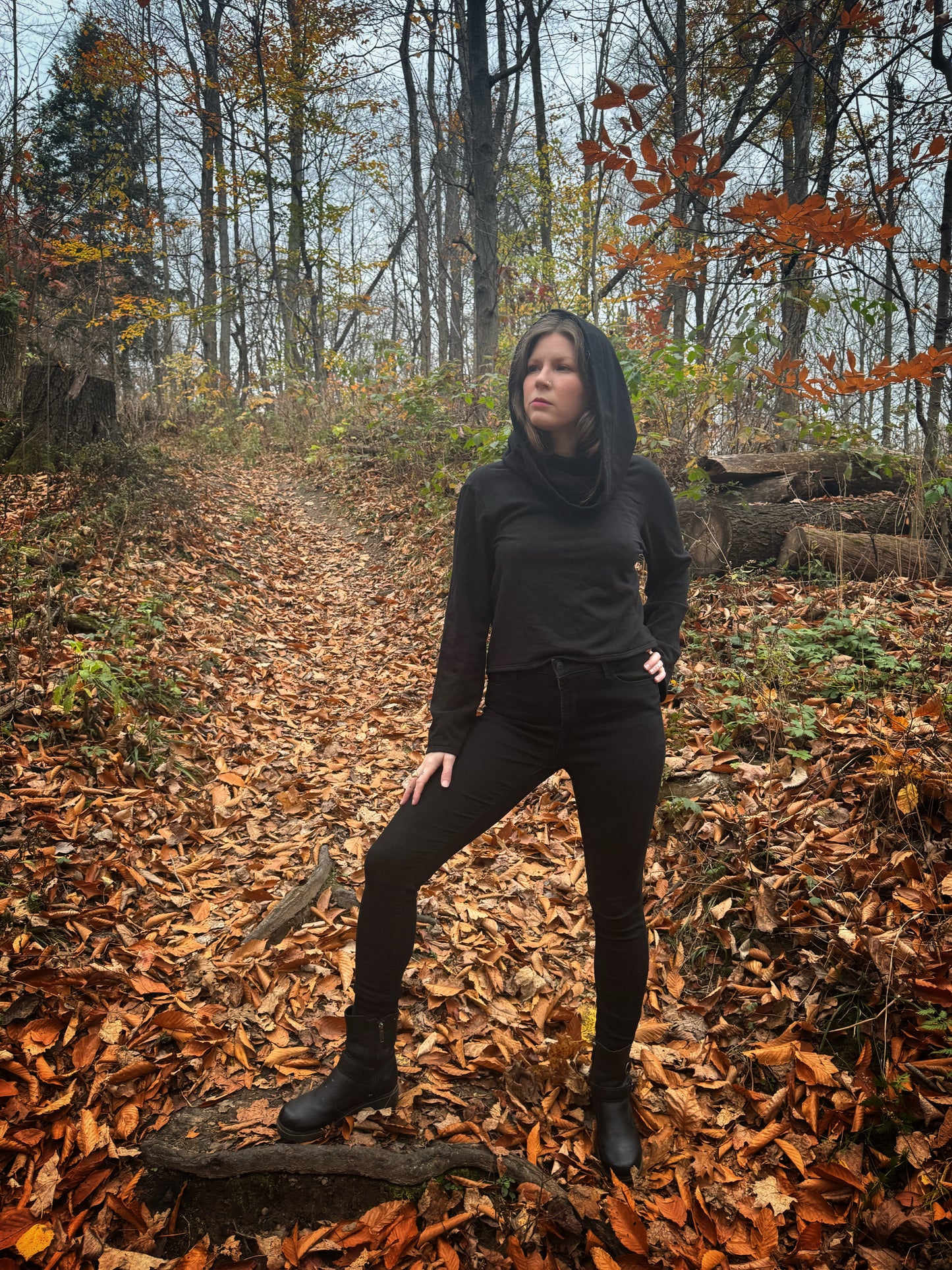 A witch standing in a fprest covered in fallen leaves wearing black pants and a black hexen hoodie. The hoodie features a large cowl hood and cropped just at top of the hips. Featuring slight bell sleeves and its made from a soft bamboo knit with a fleece interior. Very soft and warm good for layering. Witch clothing, fall wear, sustainably made in canada