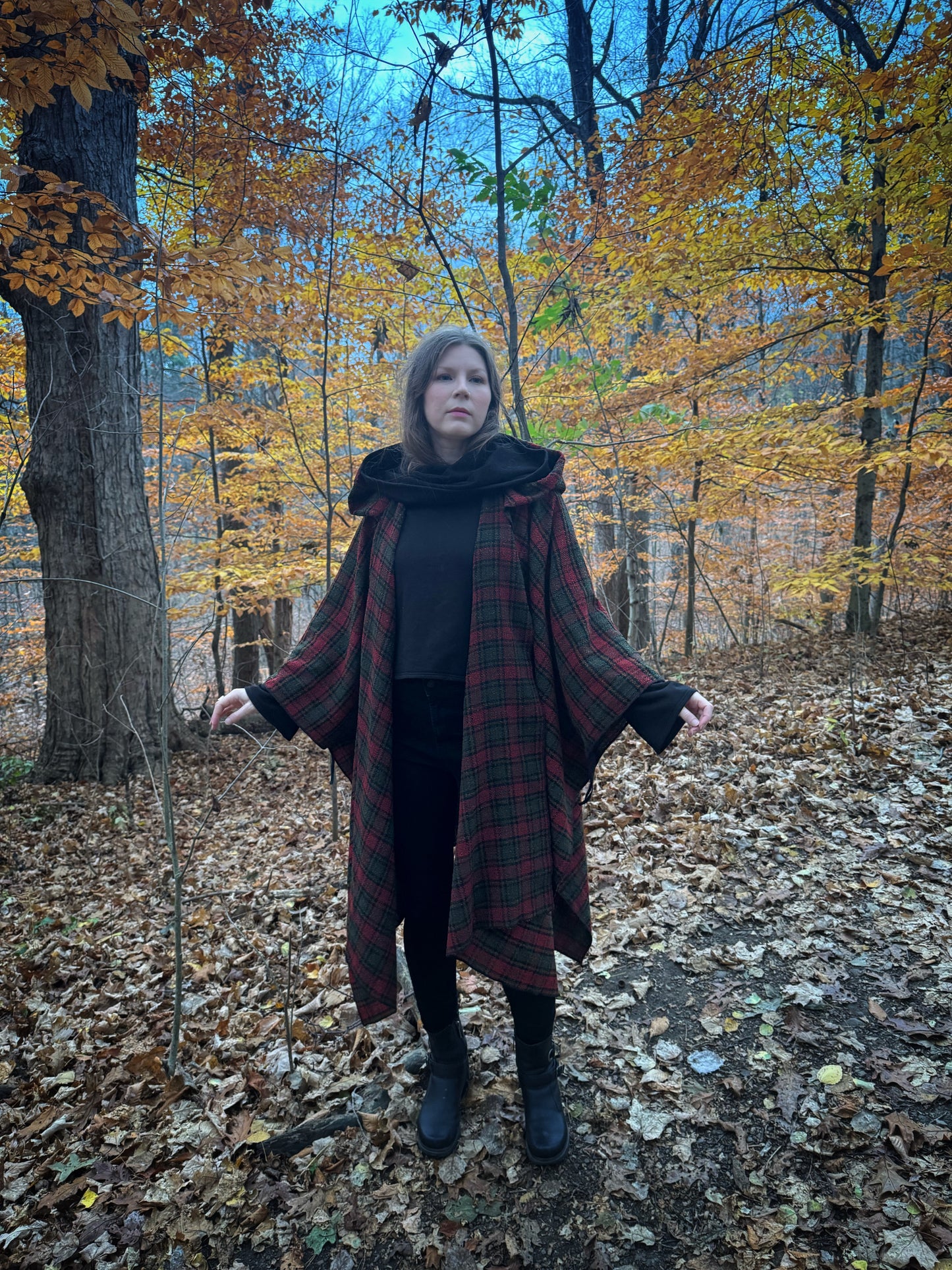 A witch standing in a forest covered in fallen leaves wearing black pants and a black hexen hoodie with a tartan poncho layered over top, the hood is down featuring the cowl. The hoodie features a large cowl hood and cropped just at top of the hips. Featuring slight bell sleeves and its made from a soft bamboo knit with a fleece interior. Very soft and warm good for layering. Witch clothing, fall wear, sustainably made in canada