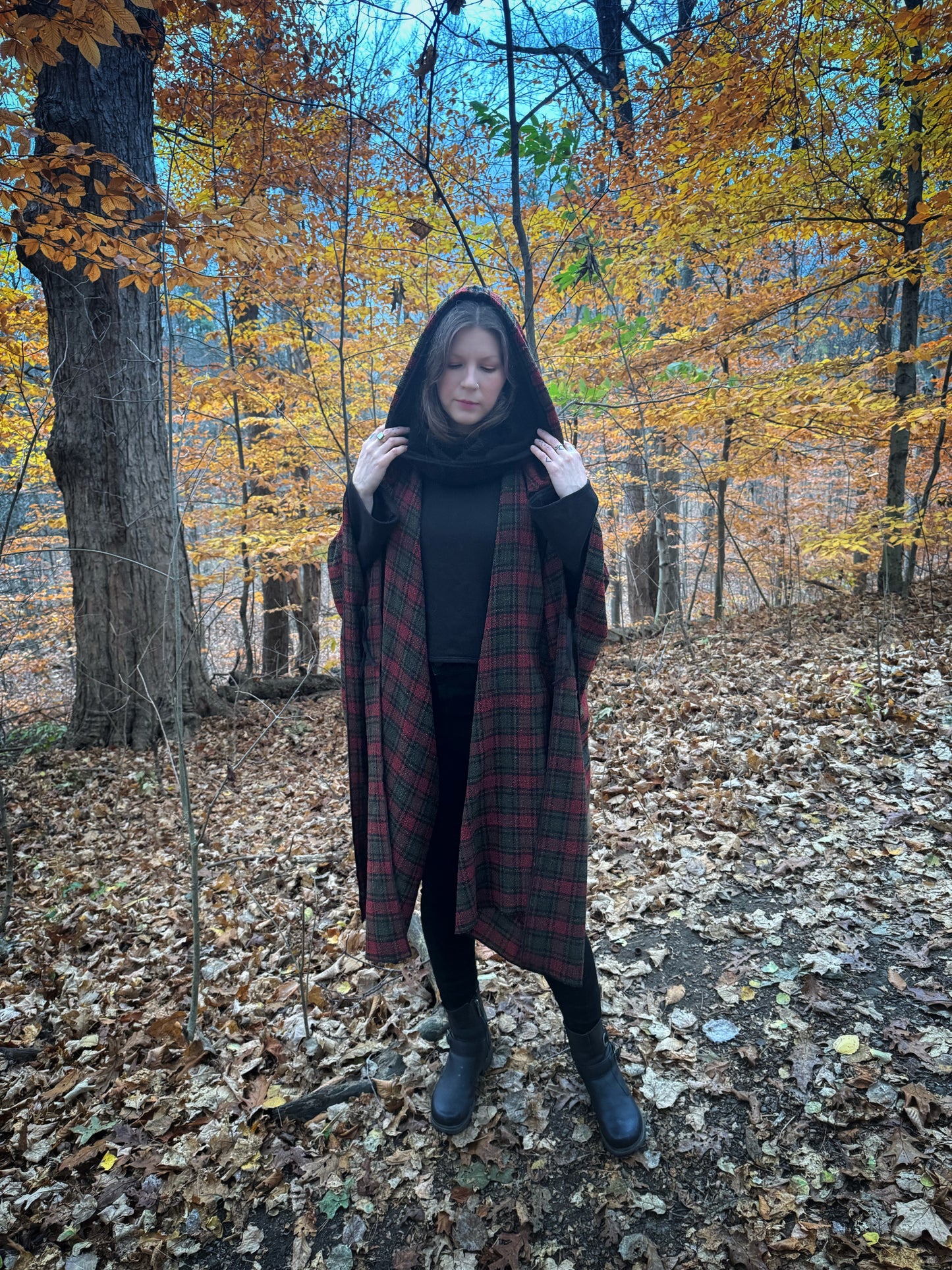 A witch standing in a fprest covered in fallen leaves wearing black pants and a black hexen hoodie with a tartan poncho layered over top. The hoodie features a large cowl hood and cropped just at top of the hips. Featuring slight bell sleeves and its made from a soft bamboo knit with a fleece interior. Very soft and warm good for layering. Witch clothing, fall wear, sustainably made in canada