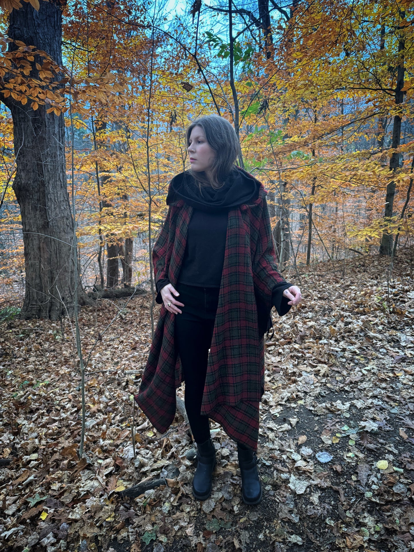 A witch standing in a forest covered in fallen leaves wearing black pants and a black hexen hoodie with a tartan poncho layered over top. The hoodie features a large cowl hood , the hood is down for this photo and cropped just at top of the hips. Featuring slight bell sleeves and its made from a soft bamboo knit with a fleece interior. Very soft and warm good for layering. Witch clothing, fall wear, sustainably made in canada