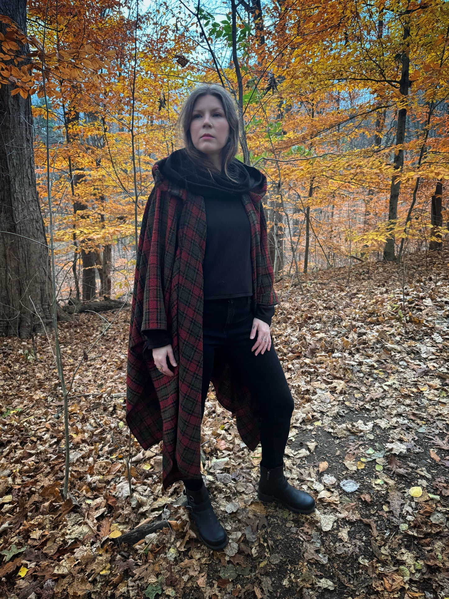 A witch standing in a forest covered in fallen leaves wearing black pants and a black hexen hoodie with a tartan poncho layered over top, the hood is down featuring the cowl. The hoodie features a large cowl hood and cropped just at top of the hips. Featuring slight bell sleeves and its made from a soft bamboo knit with a fleece interior. Very soft and warm good for layering. Witch clothing, fall wear, sustainably made in canada