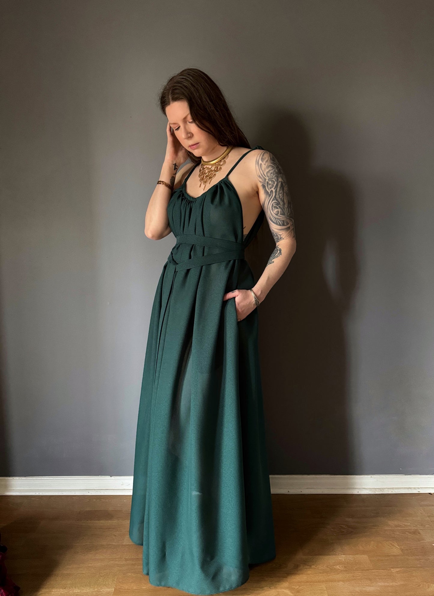 A priestess standing facing slightly to her right with her left hand in her pocket and right hand pulling back her hair. The gown is made from a forest green georgette chiffon that was upcycled deadstock. It features adjustable straps that tie at the shoulders. side seam pockets and a long wrap belt to pull in the waist. Priestess, ritual gown, witch dress, sustainable clothing, made in Canada