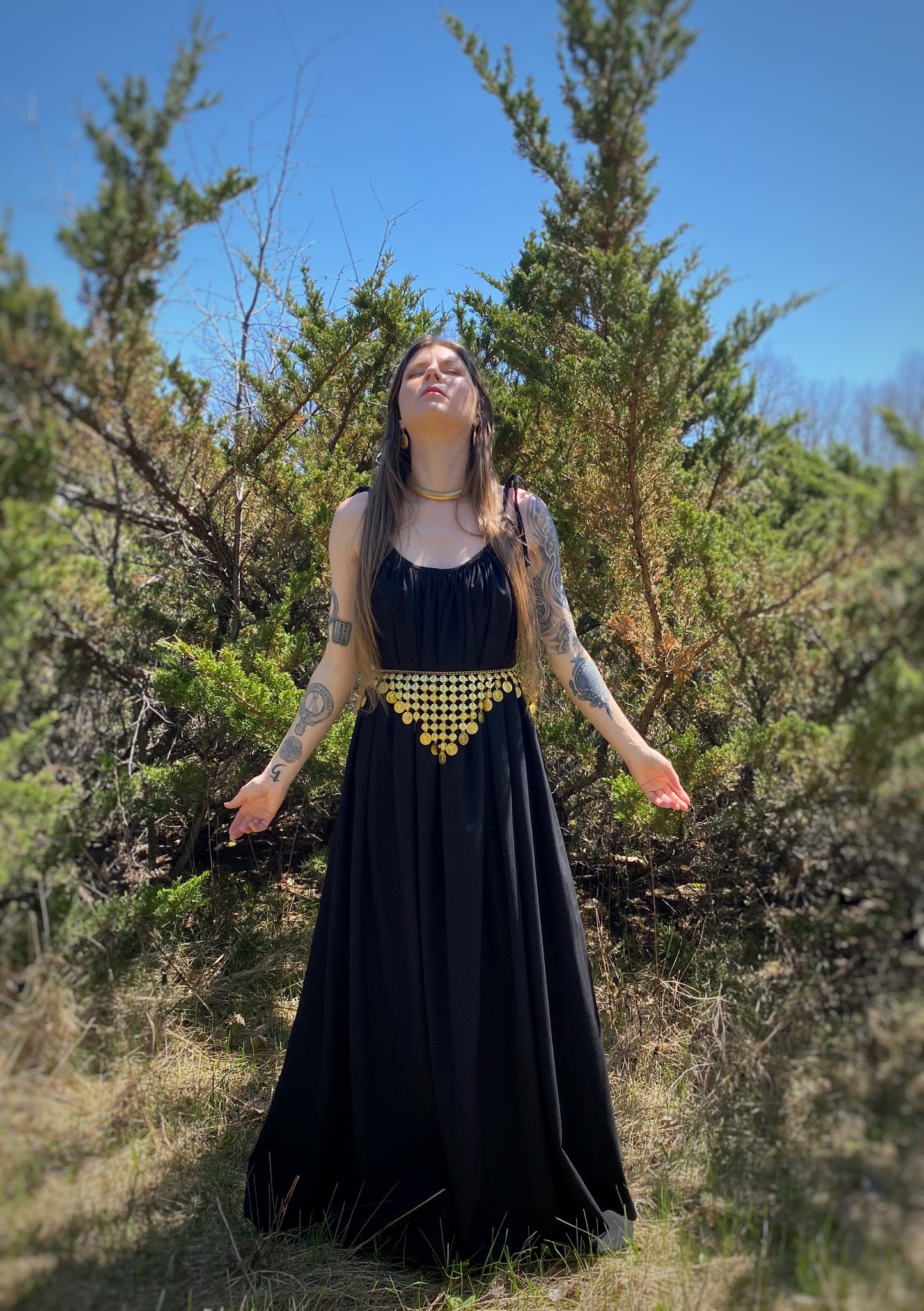 A priestess stands forward looking up with her hands out and facing forward wearing a black fates gown with a gold coin belt. The gown is made from a lack linen tencel blend, featuringajustable thin straps that tie at the shoulder, side seam pockets and comes with a long wrap belt to tie at the waist. Priestess, ritual gown. witch clothing, sustainable clothing made in canada.