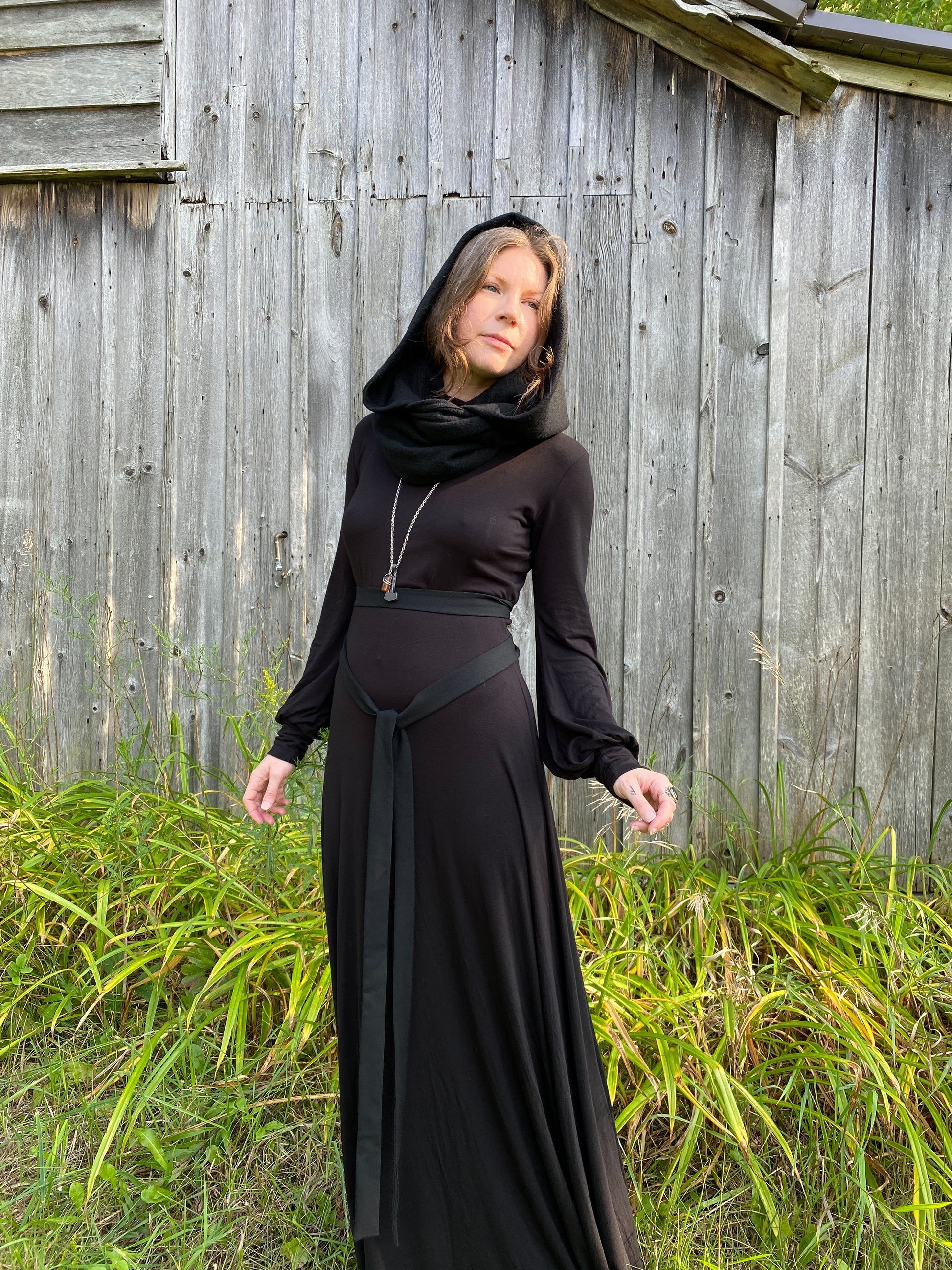 A woman with red hair in a black dress. Standing infront of a pionner, wood building with tall green across along the ground. She is showcasing a black wool cowl and hood that is being worn wrapped around the neck with the hood up.