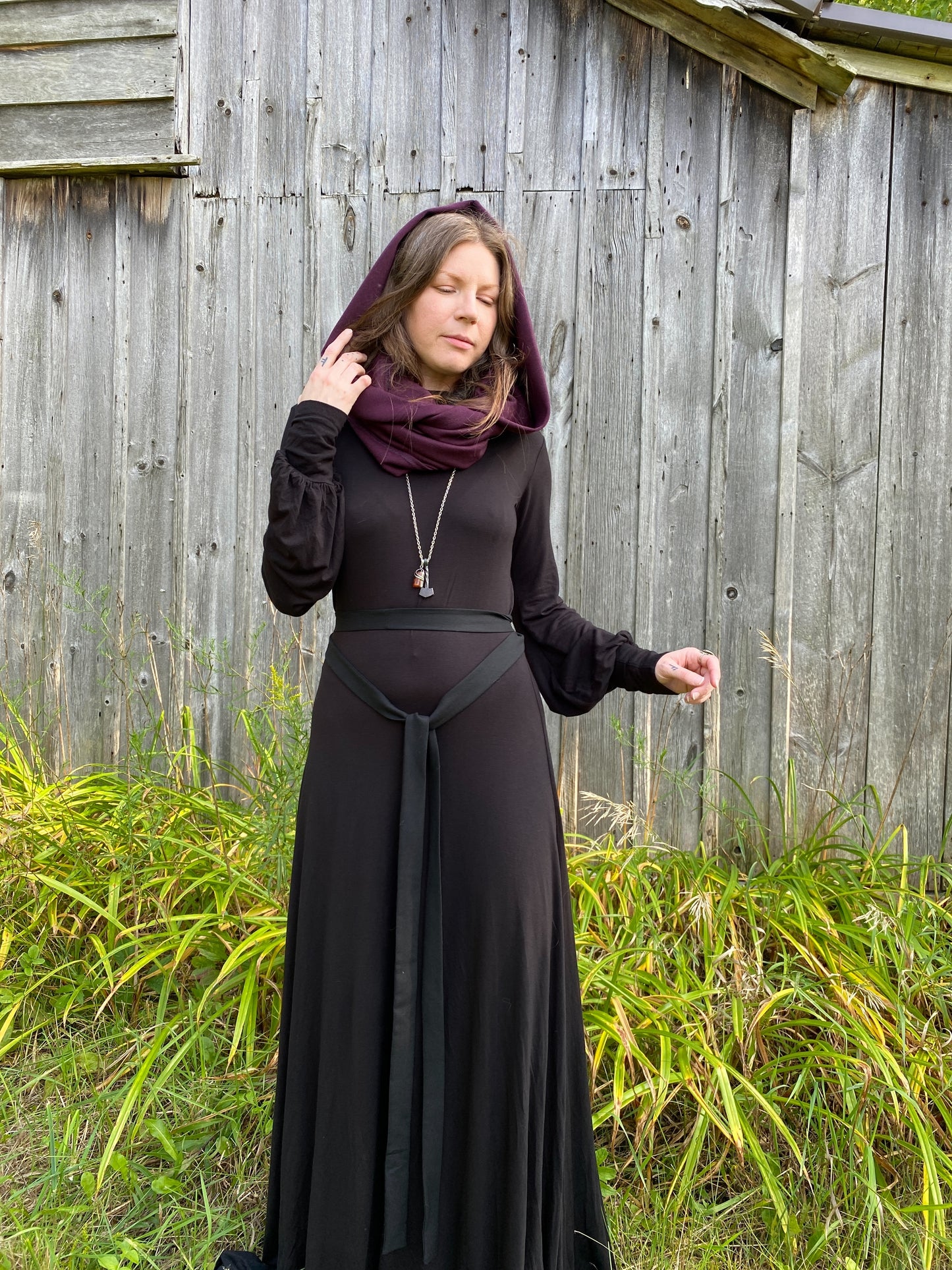 A woman with red hair in a black dress. Standing infront of a pionner, wood building with tall green across along the ground. She is showcasing a burgundy wool  cowl and hood that is being worn wrapped around the neck with the hood up.She is wearing a long silver necklace and her right hand is up, gently touching the bottom hem of the hood. 