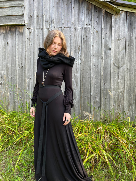 A woman with red hair in a black dress. Standing infront of a pionner, wood building with tall green across along the ground. She is showcasing a black wool cowl and hood that is being worn wrapped around the neck with the hood down.