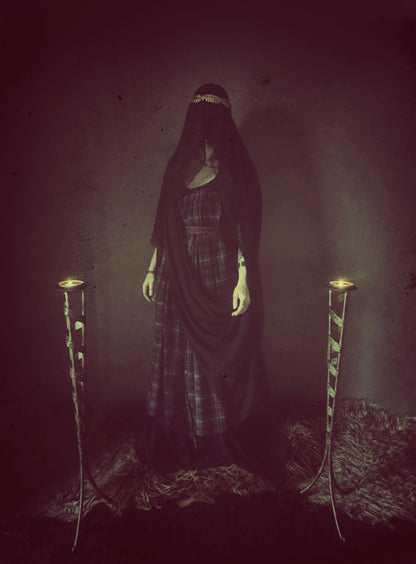 A high priestess stands between two flames. Wearing a tartan dress with a long black veiled wrapped around her body and cover her face. The plaid dress is floor length with adjustable thin shoulder straps featuring side seam pockets and a long thin black wrap belt. The veil I black made from recycled chiffon. 