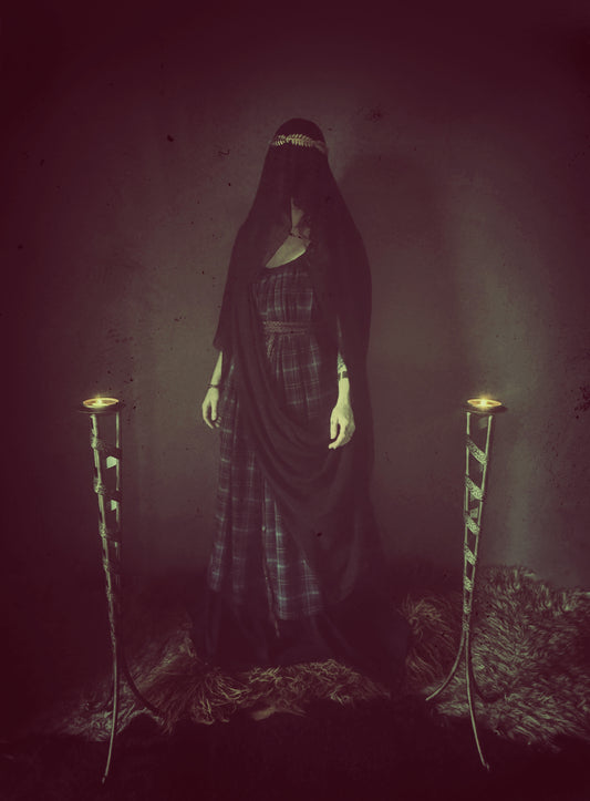 A high priestess stands between two flames. Wearing a tartan dress with a long black veiled wrapped around her body and cover her face. The plaid dress is floor length with adjustable thin shoulder straps featuring side seam pockets and a long thin black wrap belt. The veil I black made from recycled chiffon. 