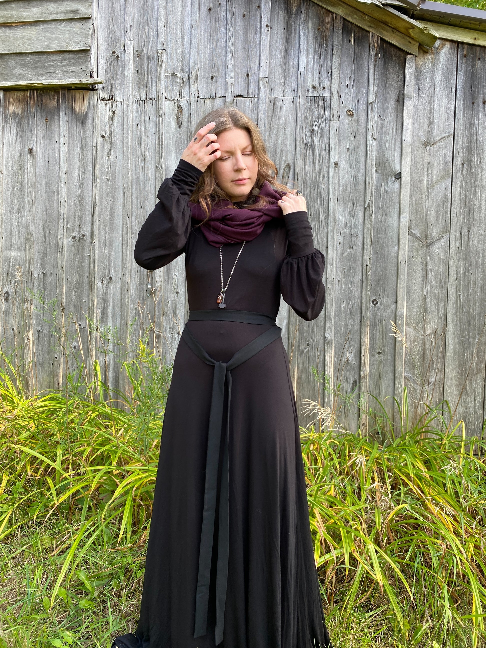 A woman with red hair in a black dress. Standing infront of a pionner, wood building with tall green across along the ground. She is showcasing a burgundy wool cowl and hood that is being worn wrapped around the neck with the hood up.She is wearing a long silver necklace and her right hand is up, gently touching the bottom hem of the hood. Her right hand is up gently pulling back her hair from her face.