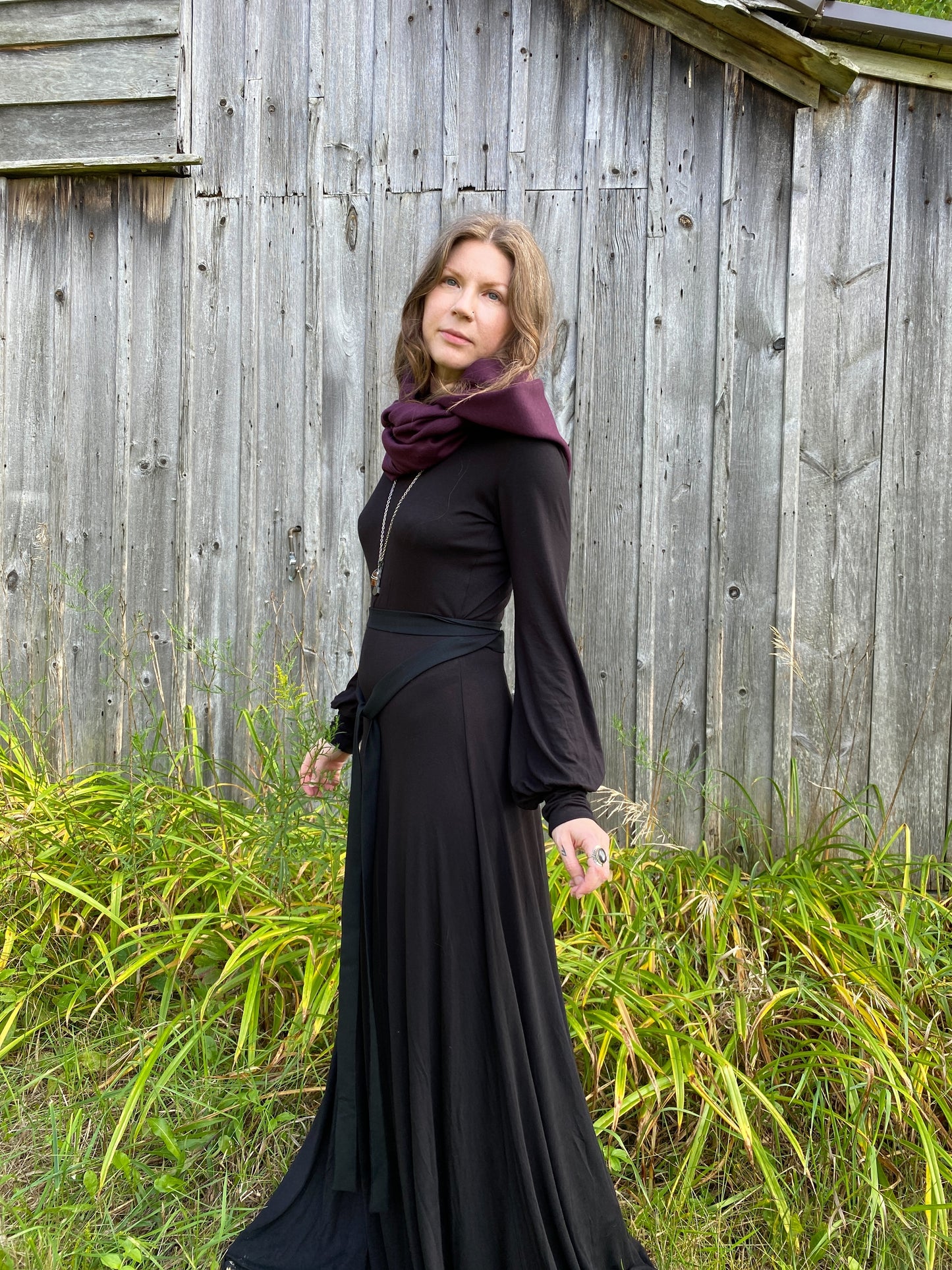 A woman with red hair in a black dress. Standing infront of a pionner, wood building with tall green across along the ground. She is showcasing a burgundy wool cowl and hood that is being worn wrapped around the neck with the hood down. 