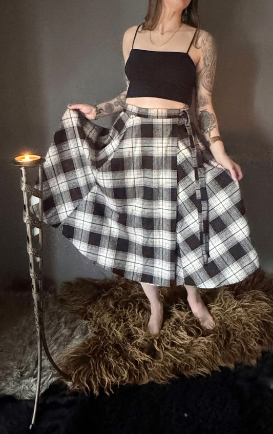 A photo of a black and white plaid wrap skirt. Only the lower half the body is showing featuring the mid calf length and long ties. An adjustable cotton tartan wrap skirt, size inclusive and made in Canada.
