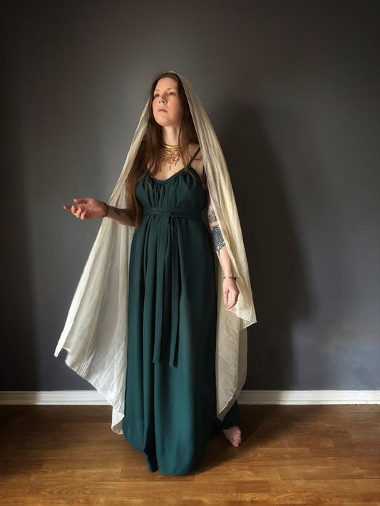 A woman standing forward with her right arm reached out. Wearing a forest green fates gown. featuring adjustable thin straps that tie at the shoulder, side seam pockets and a long wrap belt at the waist. She is also wearing our cotton gauze priestess veil draped over her head. Priestess, ritual gown, witch clothing, living goddess. 