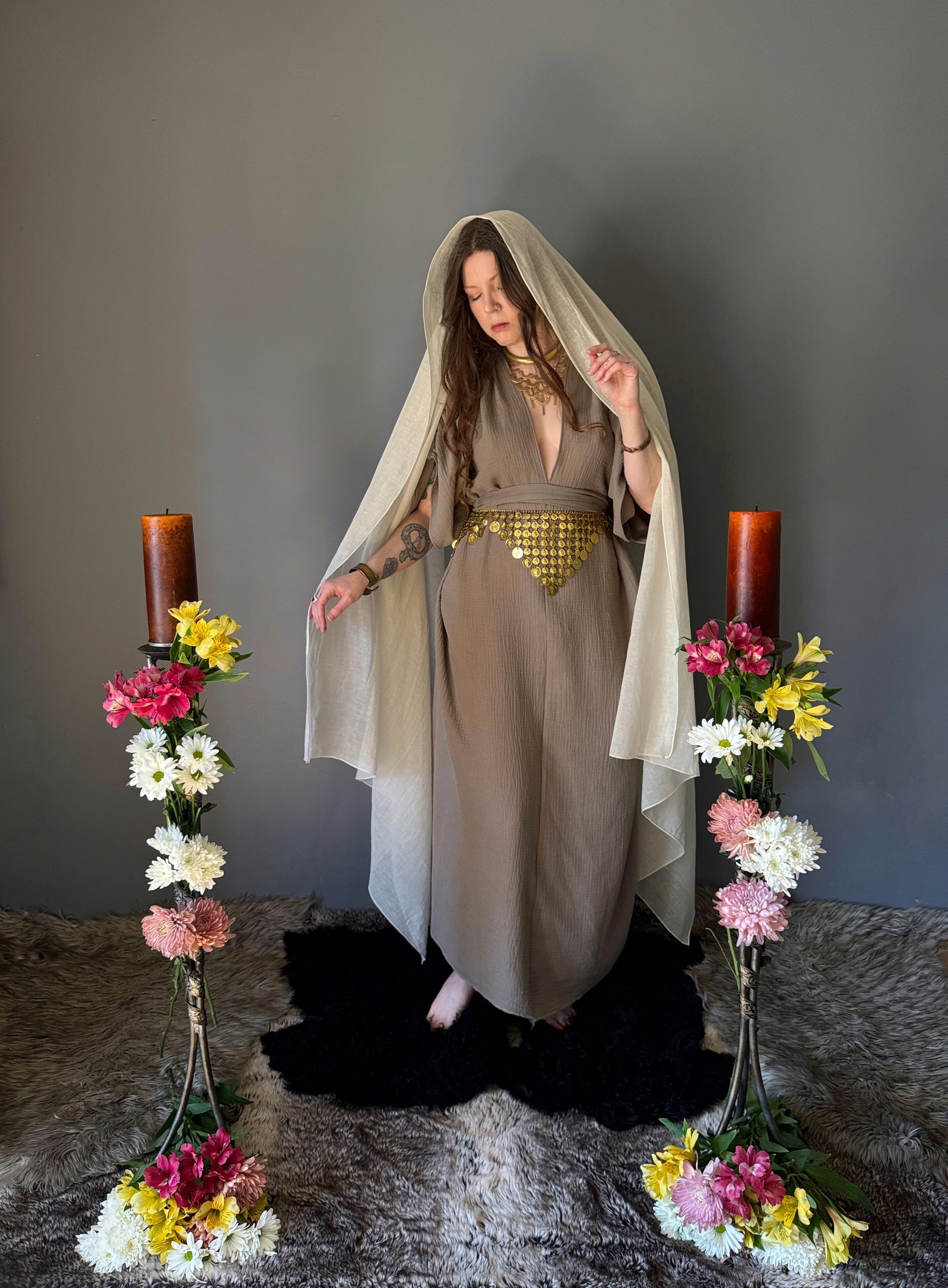 A priestess surrounded by flowers wearing a ancient greek inspired ritual robe. Featuring a deep v neck line, gathered short sleeves with braided trim that ties at the back. Cinched at the waist with a matching long wrap belt, draping the veil over her head. The gown is a tan colour cotton with a slight waffle comb texture. The veil is made from a cotton gauze in a natural undyed state. For the magically inclined, witches, priestess, priest, pagan, hellenistic, polytheistic, spiritual or lover of history.