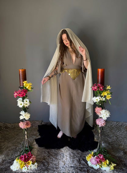 A priestess surrounded by flowers wearing a ancient greek inspired ritual robe. Featuring a deep v neck line, gathered short sleeves with braided trim that ties at the back. Cinched at the waist with a matching long wrap belt, draping the veil over her head. The gown is a tan colour cotton with a slight waffle comb texture. The veil is made from a cotton gauze in a natural undyed state. For the magically inclined, witches, priestess, priest, pagan, hellenistic, polytheistic, spiritual or lover of history.