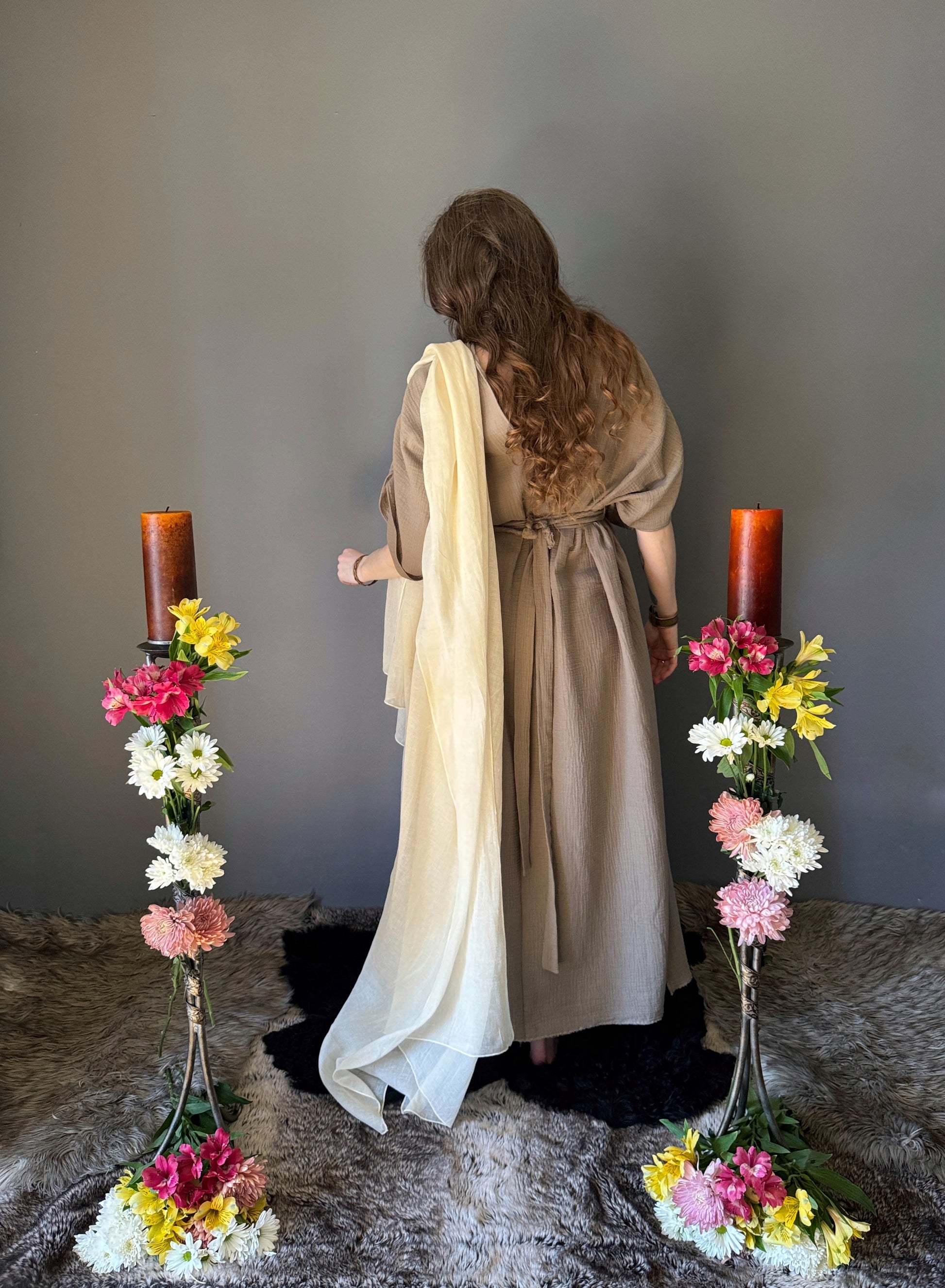 A priestess surrounded by flowers wearing a ancient greek inspired ritual robe. Showcasing the back, gathered short sleeves with braided trim that ties at the back neck and veil draped over shoulder. Cinched at the waist with a matching long wrap belt. The gown is a soft tan colour cotton with a slight waffle comb texture. The veil is made from a cotton gauze in a natural undyed state. For the magically inclined, witches, priestess, priest, pagan, hellenistic, polytheistic, spiritual or lover of history.