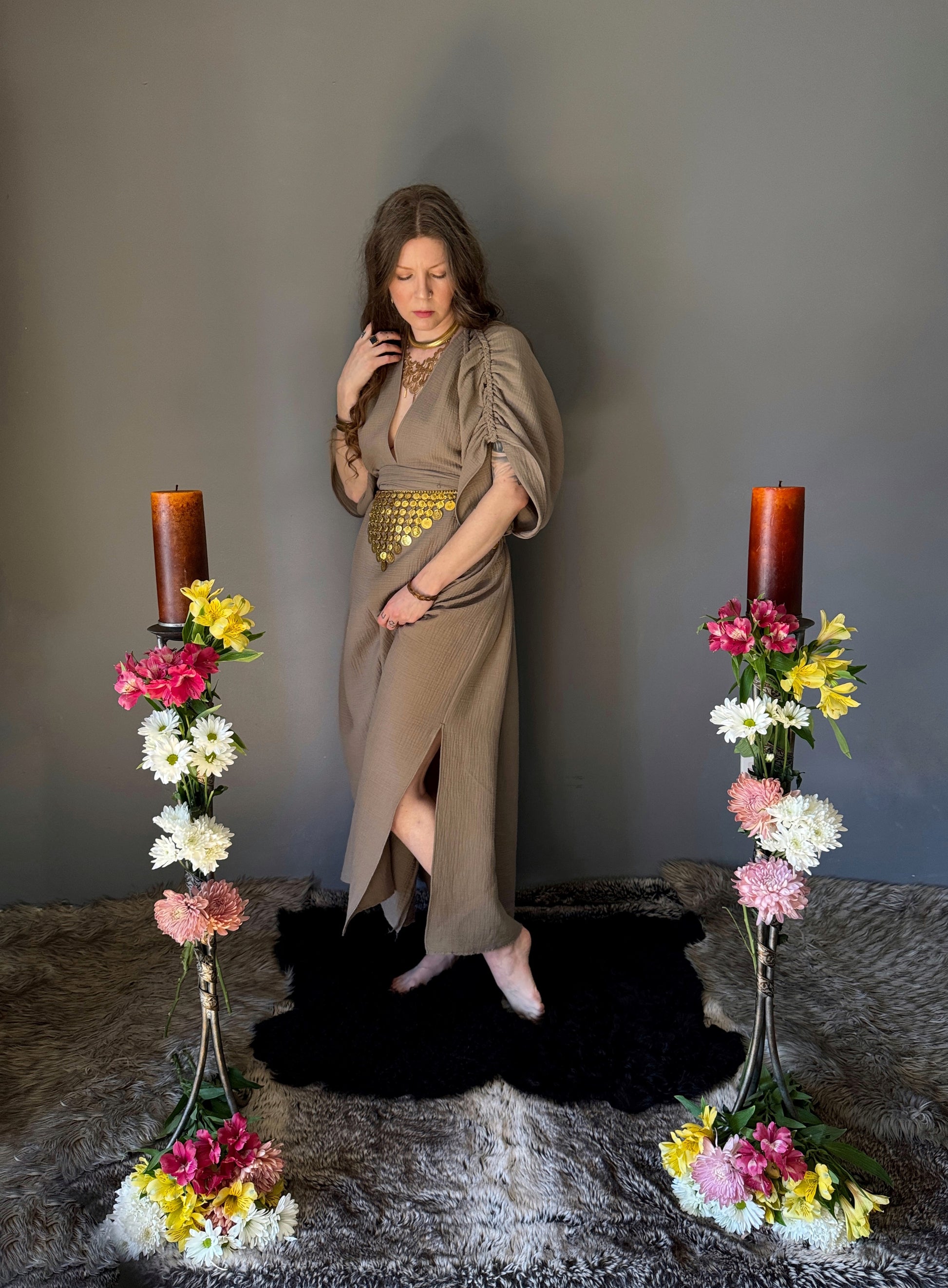 A priestess surrounded by flowers wearing a ancient greek inspired ritual robe. Featuring a deep v neck line, gathered short sleeves with braided trim that ties at the back. Cinched at the waist with a matching long wrap belt. Showcasing the roughly knee height side slits. The gown is a soft tan colour cotton with a slight waffle comb texture. For the magically inclined, witches, priestess, priest, pagan, hellenistic, polytheistic, spiritual or lover of history.