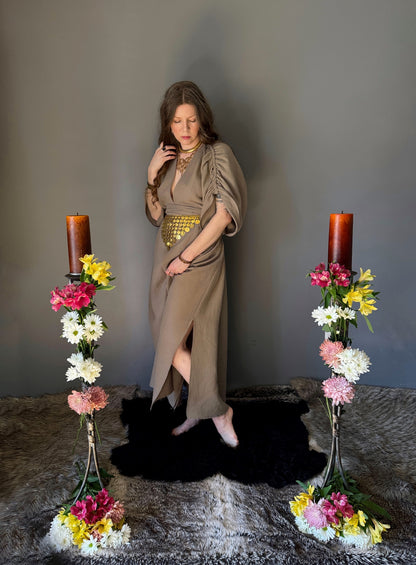 A priestess surrounded by flowers wearing a ancient greek inspired ritual robe. Featuring a deep v neck line, gathered short sleeves with braided trim that ties at the back. Cinched at the waist with a matching long wrap belt. Showcasing the roughly knee height side slits. The gown is a soft tan colour cotton with a slight waffle comb texture. For the magically inclined, witches, priestess, priest, pagan, hellenistic, polytheistic, spiritual or lover of history.
