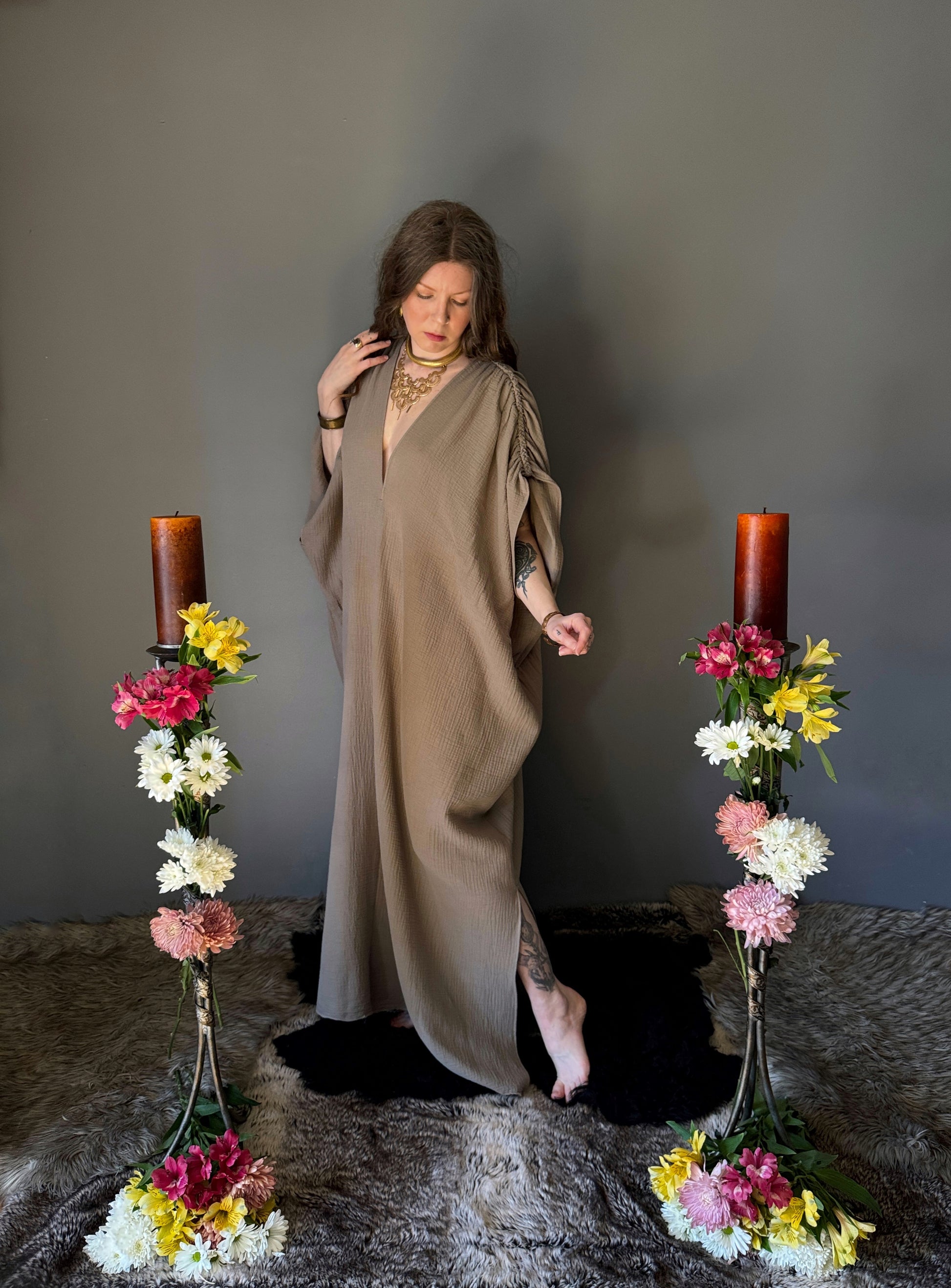 A priestess surrounded by flowers wearing a ancient greek inspired ritual robe. Featuring a deep v neck line, gathered short sleeves with braided trim that ties at the back. Cinched at the waist with a matching long wrap belt. The gown is a soft tan colour cotton with a slight waffle comb texture. The veil is made from a cotton gauze in a natural undyed state. For the magically inclined, witches, priestess, priest, pagan, hellenistic, polytheistic, spiritual or lover of history.No belt or veil in this photo