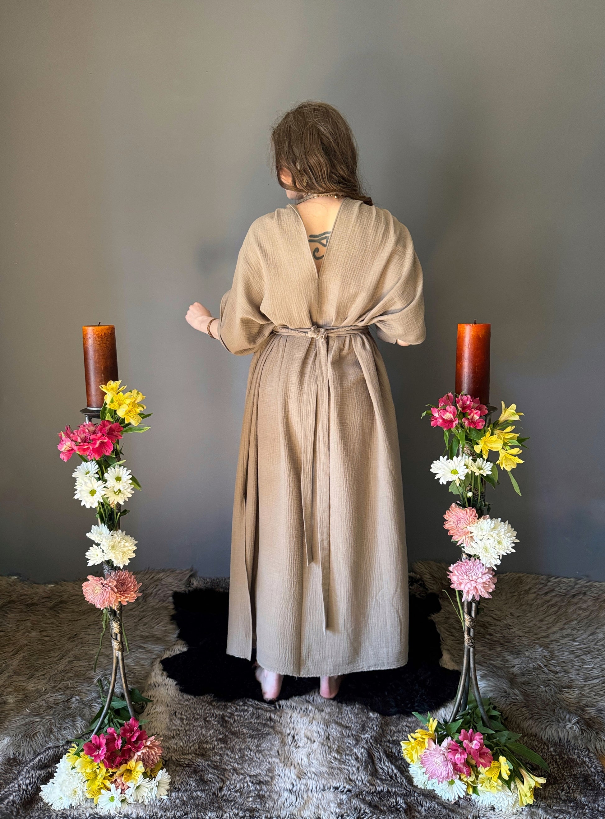 A priestess surrounded by flowers wearing a ancient greek inspired ritual robe. showing the back view with a shorter v neckline that can be reversed to the front neckline, gathered short sleeves with braided trim that ties at the back. Cinched at the waist with a matching long wrap belt. The gown is a soft tan colour cotton with a slight waffle comb texture. For the magically inclined, witches, priestess, priest, pagan, hellenistic, polytheistic, spiritual or lover of history.