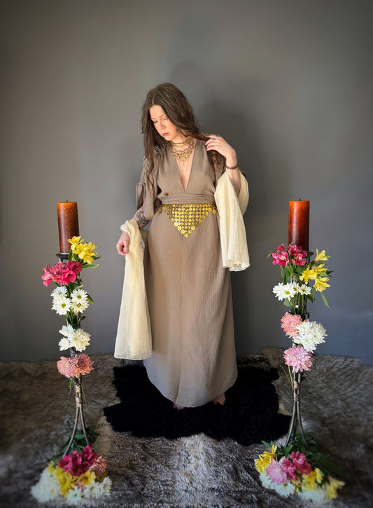 A priestess surrounded by flowers wearing a ancient greek inspired ritual robe. Featuring a deep v neck line, gathered short sleeves with braided trim that ties at the back. Cinched at the waist with a matching long wrap belt. The gown is a soft tan colour cotton with a slight waffle comb texture. The veil is made from a cotton gauze in a natural undyed state. For the magically inclined, witches, priestess, priest, pagan, hellenistic, polytheistic, spiritual or lover of history.