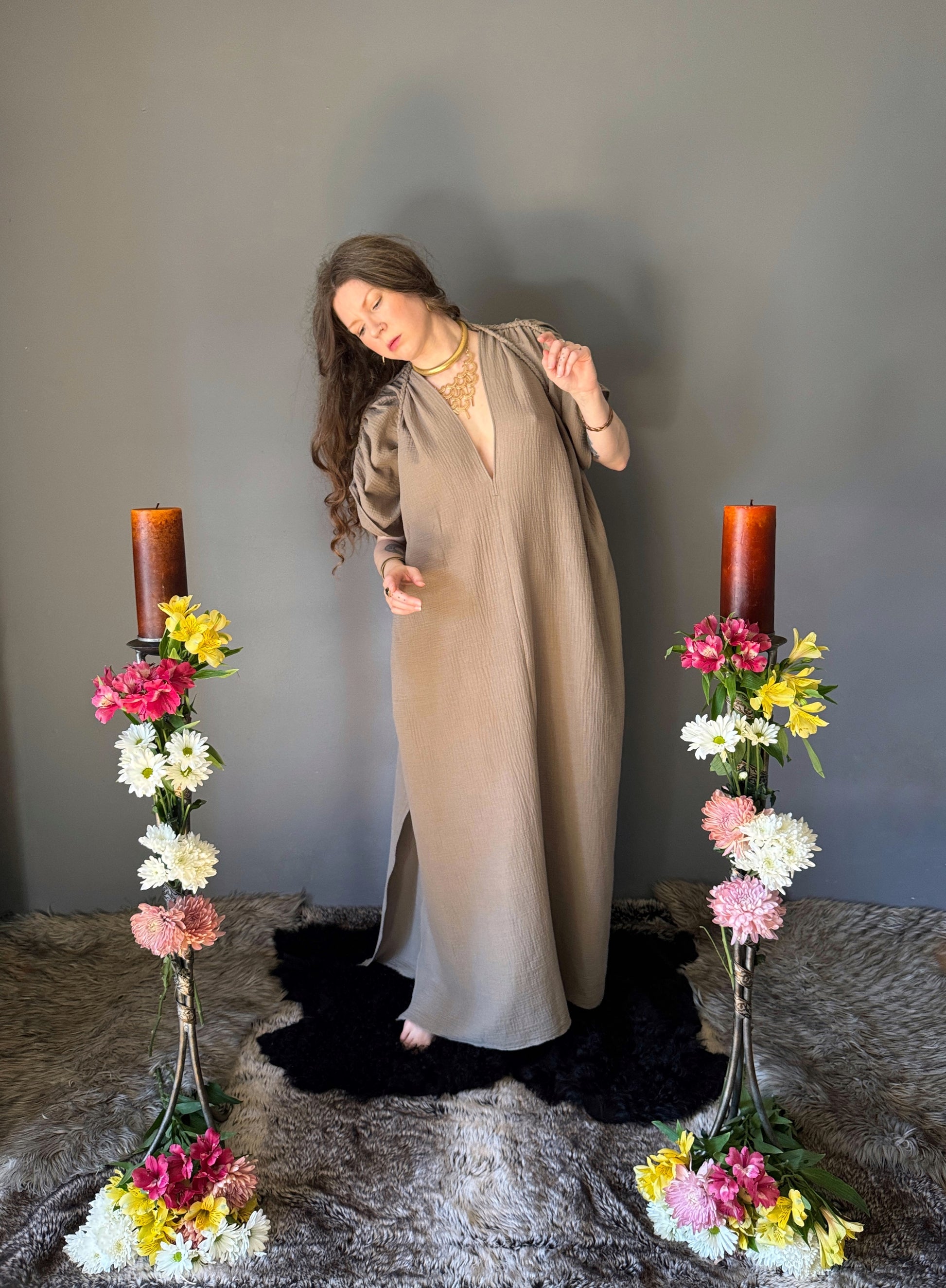 A priestess surrounded by flowers wearing a ancient greek inspired ritual robe. Featuring a deep v neck line, gathered short sleeves with braided trim that ties at the back. The gown is a soft tan colour cotton with a slight waffle comb texture. For the magically inclined, witches, priestess, priest, pagan, hellenistic, polytheistic, spiritual or lover of history.