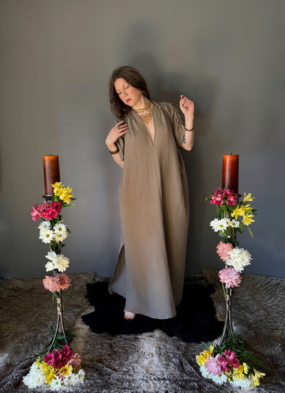 A priestess surrounded by flowers wearing a ancient greek inspired ritual robe. Featuring a deep v neck line, gathered short sleeves with braided trim that ties at the back.  The gown is a soft tan colour cotton with a slight waffle comb texture. For the magically inclined, witches, priestess, priest, pagan, hellenistic, polytheistic, spiritual or lover of history.