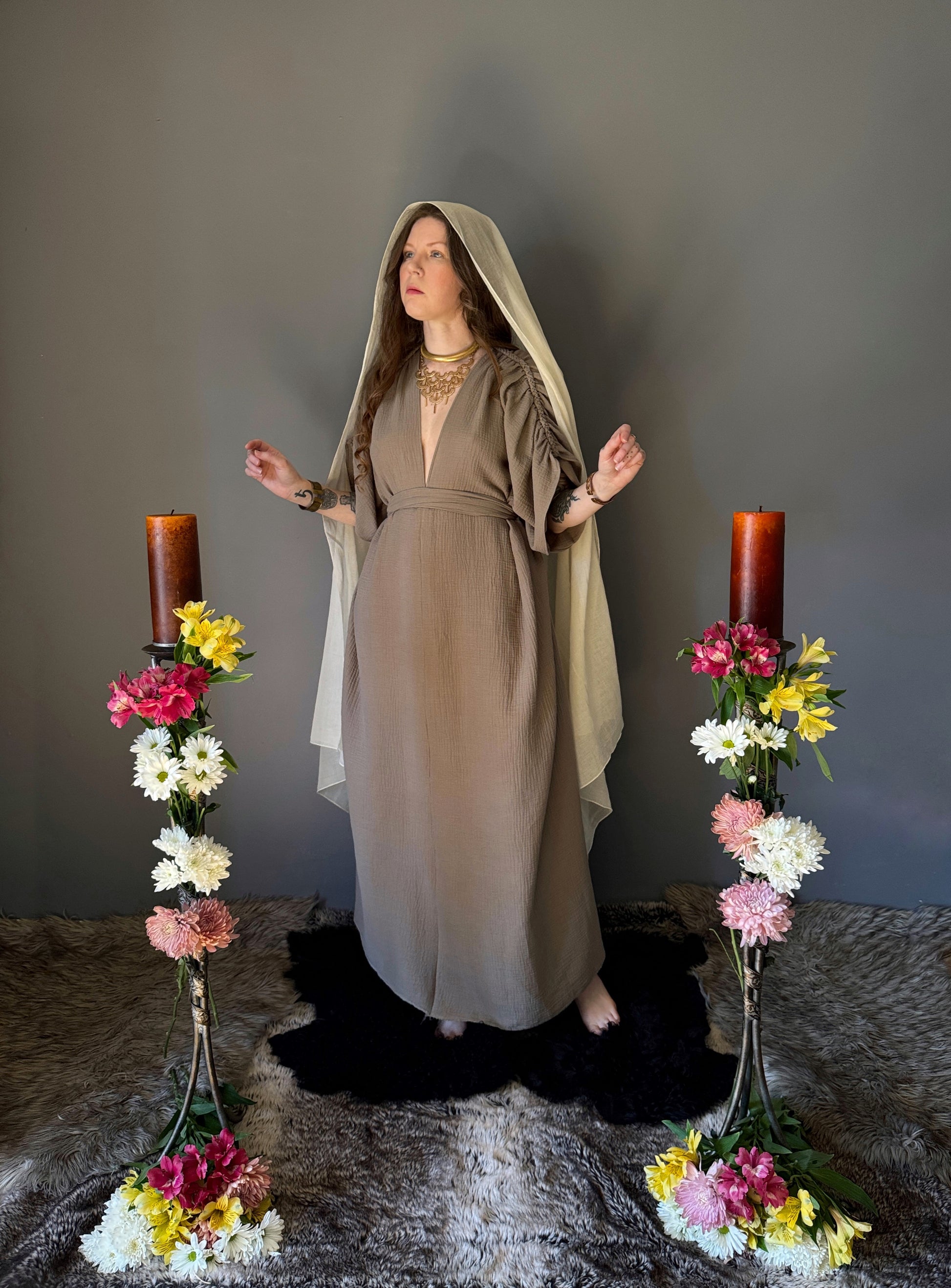 A priestess surrounded by flowers wearing a ancient greek inspired ritual robe. Featuring a deep v neck line, gathered short sleeves with braided trim that ties at the back. Cinched at the waist with a matching long wrap belt. The gown is a soft tan colour cotton with a slight waffle comb texture. The veil is made from a cotton gauze in a natural undyed state. For the magically inclined, witches, priestess, priest, pagan, hellenistic, polytheistic, spiritual or lover of history. Sustainably made in Canada