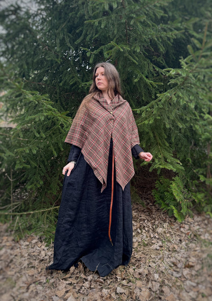 A celtic woman stands in front  some evergreen trees wearing a black tunic dress with a brown long belt and featuring an earth toned plaid shawl. Brown undertones with white and red stripes. In this photo the shawl is draped over the shoulders and held together at the front with a brass swirl cloak pin that comes with the shawl. Historical costume, ancestor reverence, old gods, made in canada.