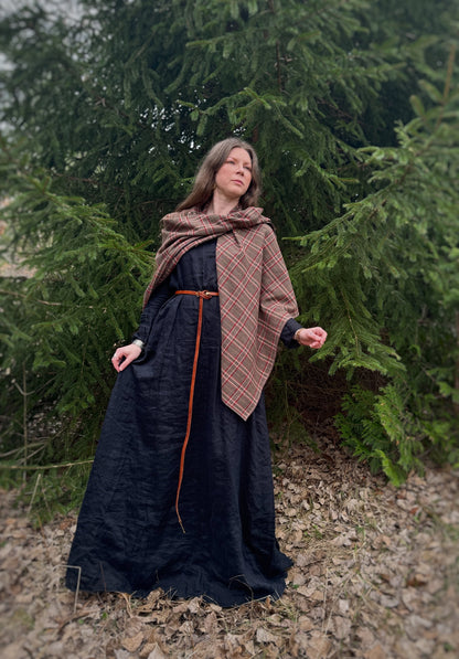 A celtic woman stands in front  some evergreen trees wearing a black tunic dress with a brown long belt and featuring an earth toned plaid shawl. Brown undertones with white and red stripes. In this photo it is a side front view with the right half of the shawl pinned up on the left side of the shoulder. Using the brass swirl cloak pin.Historical costume, ancestor reverence, old gods, made in anada.