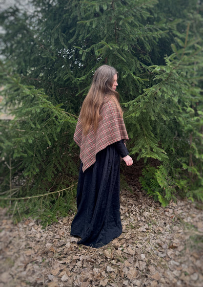 A celtic woman stands in front  some evergreen trees wearing a black tunic dress with a brown long belt and featuring an earth toned plaid shawl. Brown undertones with white and red stripes. in this photo it is a back side view of the shawl draped over the shoulders and closed at the front. You can see the point of the shawl and size. Historical costume, ancestor reverence, old gods, made in canada.