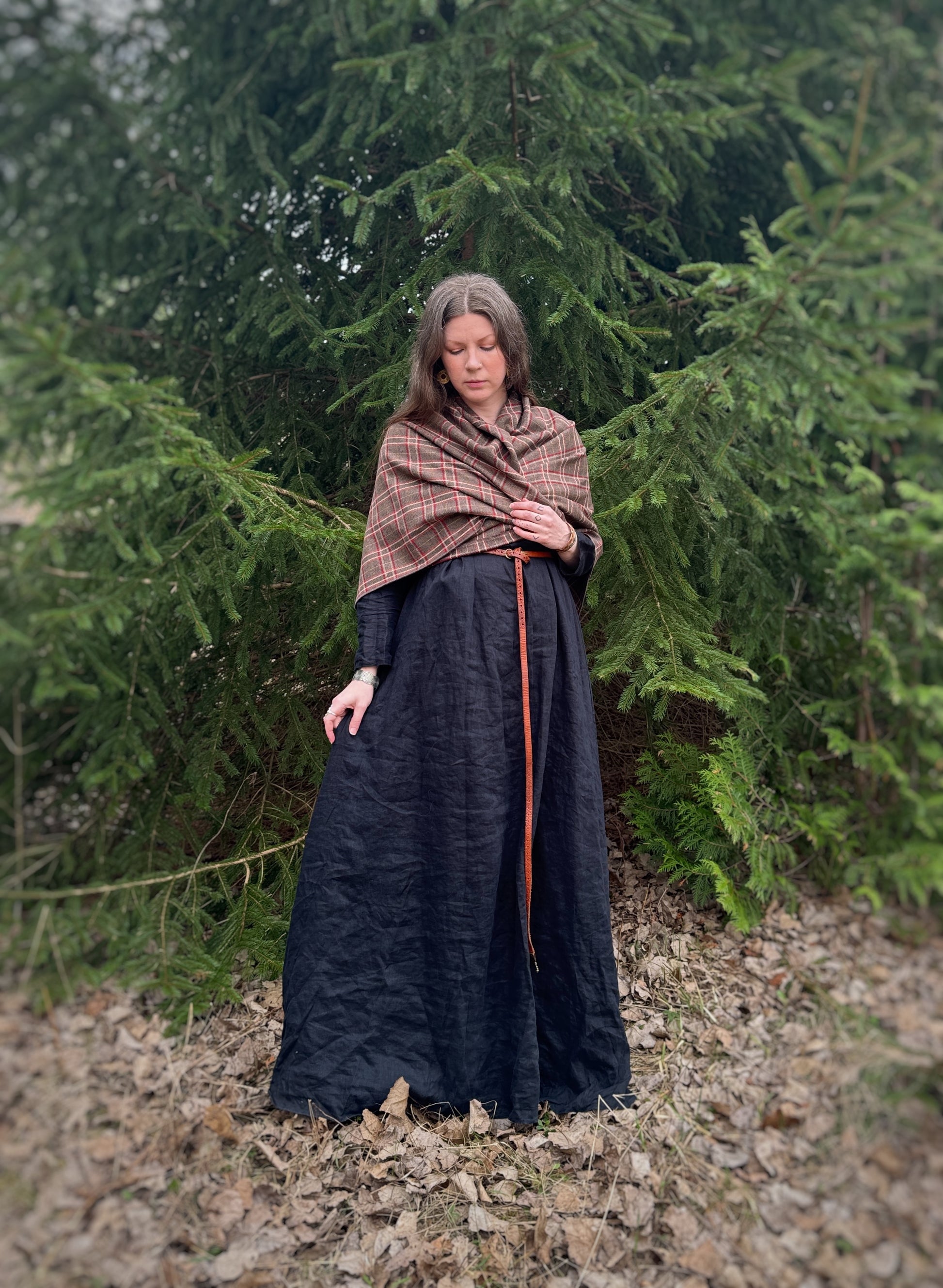 A celtic woman stands in front  some evergreen trees wearing a black tunic dress with a brown long belt and featuring an earth toned plaid shawl. Brown undertones with white and red stripes. Draped over the shoulders and crossed over the fron and tied in the back for a warm and comfortable look and feel in this photo.
Historical costume, ancestor reverence, old gods, made in canada.