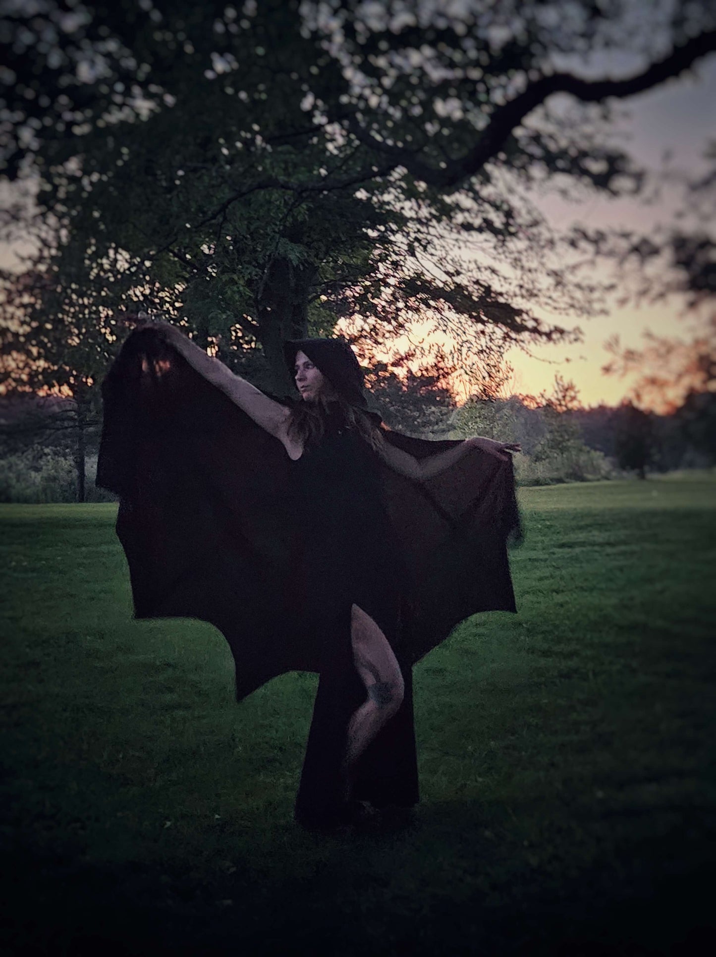 A black lightweight bat wing shaped cape. This photo is in the sunset with the cape being held up in the light by the finger loops to  showcase the bat wing hem.