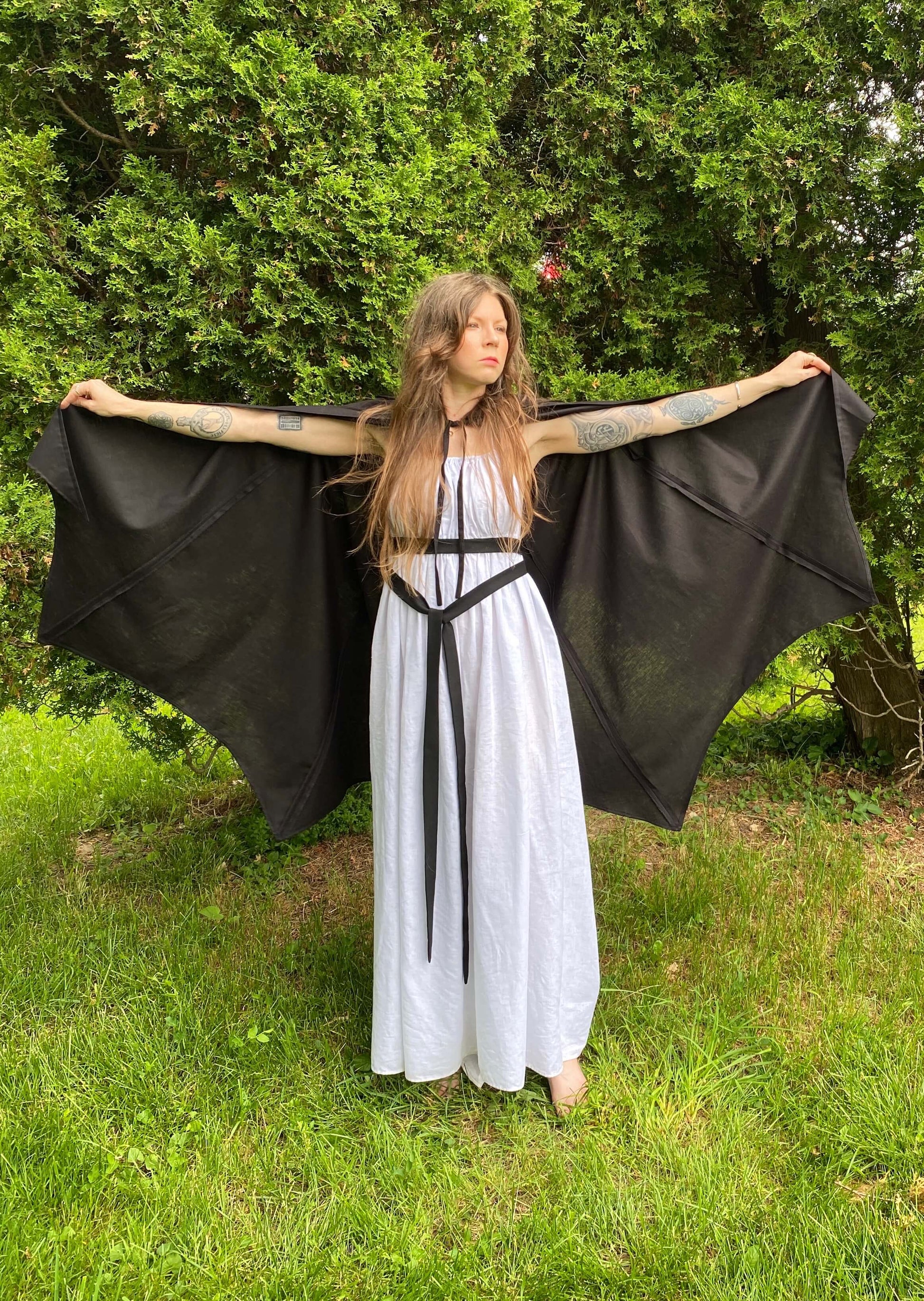 A black, lightweight bat wing shaped cape. The model is holding the cape open by the finger loops to expose the bat wing hem. 