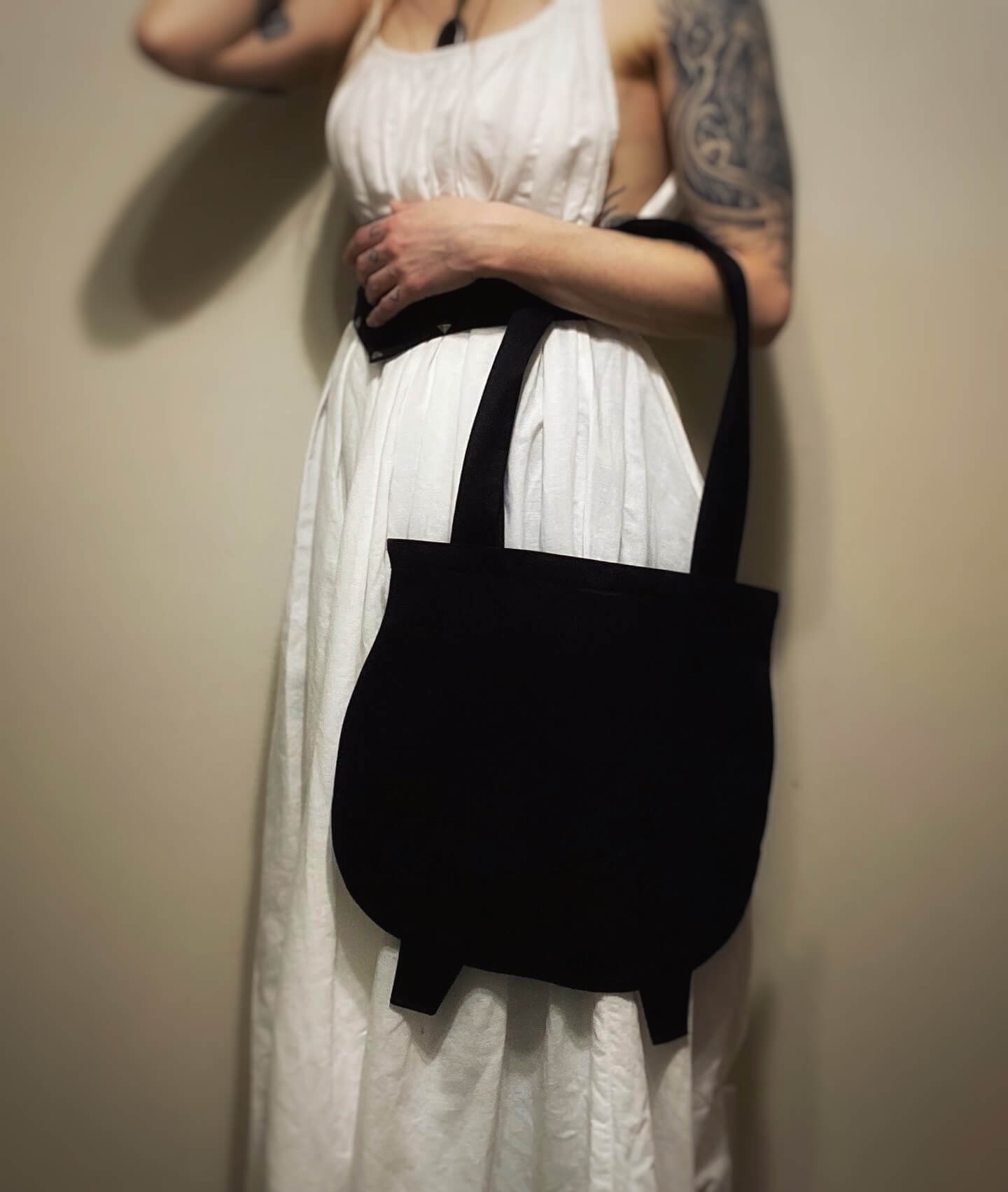This phot showcases a black tote bag in the shape of a witches cauldron. Its resting on a woman's left forearm. She is wearing a white dress, only her torso to is visible in the photo.