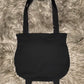 A black tote bag in the shape of a witches cauldron. With two little cauldron feet on the bottom and two shoulder straps.