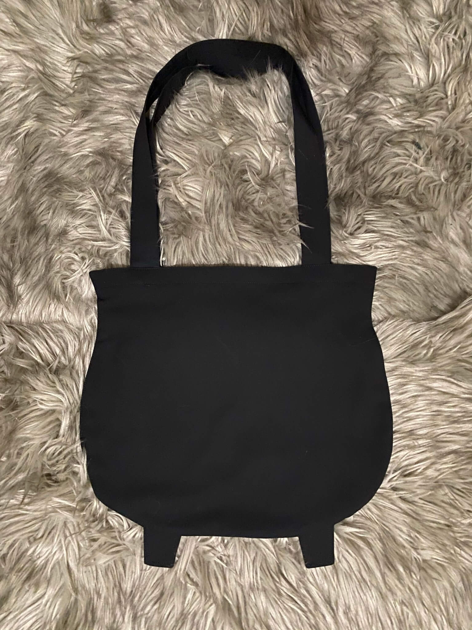 A black tote bag in the shape of a witches cauldron. With two little cauldron feet on the bottom and two shoulder straps.