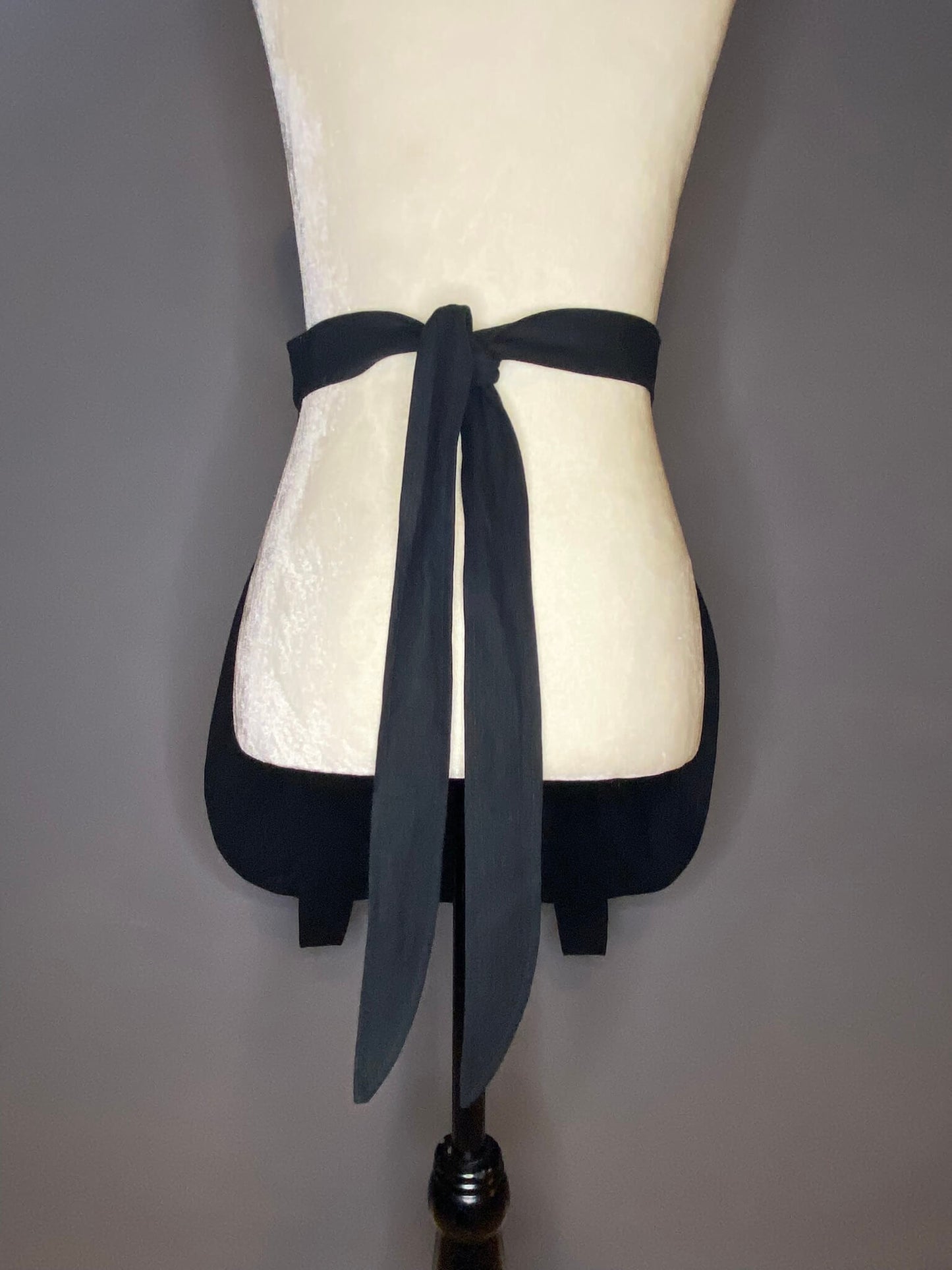 back view of a black, lined cotton linen apron in the shape of a cauldron. Back features two long ties for either your waist or hips.