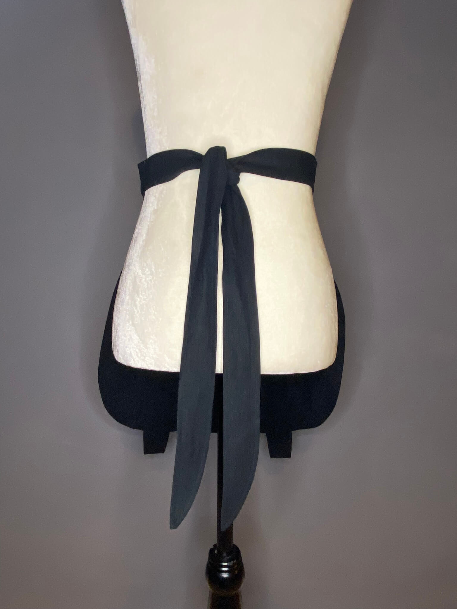 back view of a black, lined cotton linen apron in the shape of a cauldron. Back features two long ties for either your waist or hips.