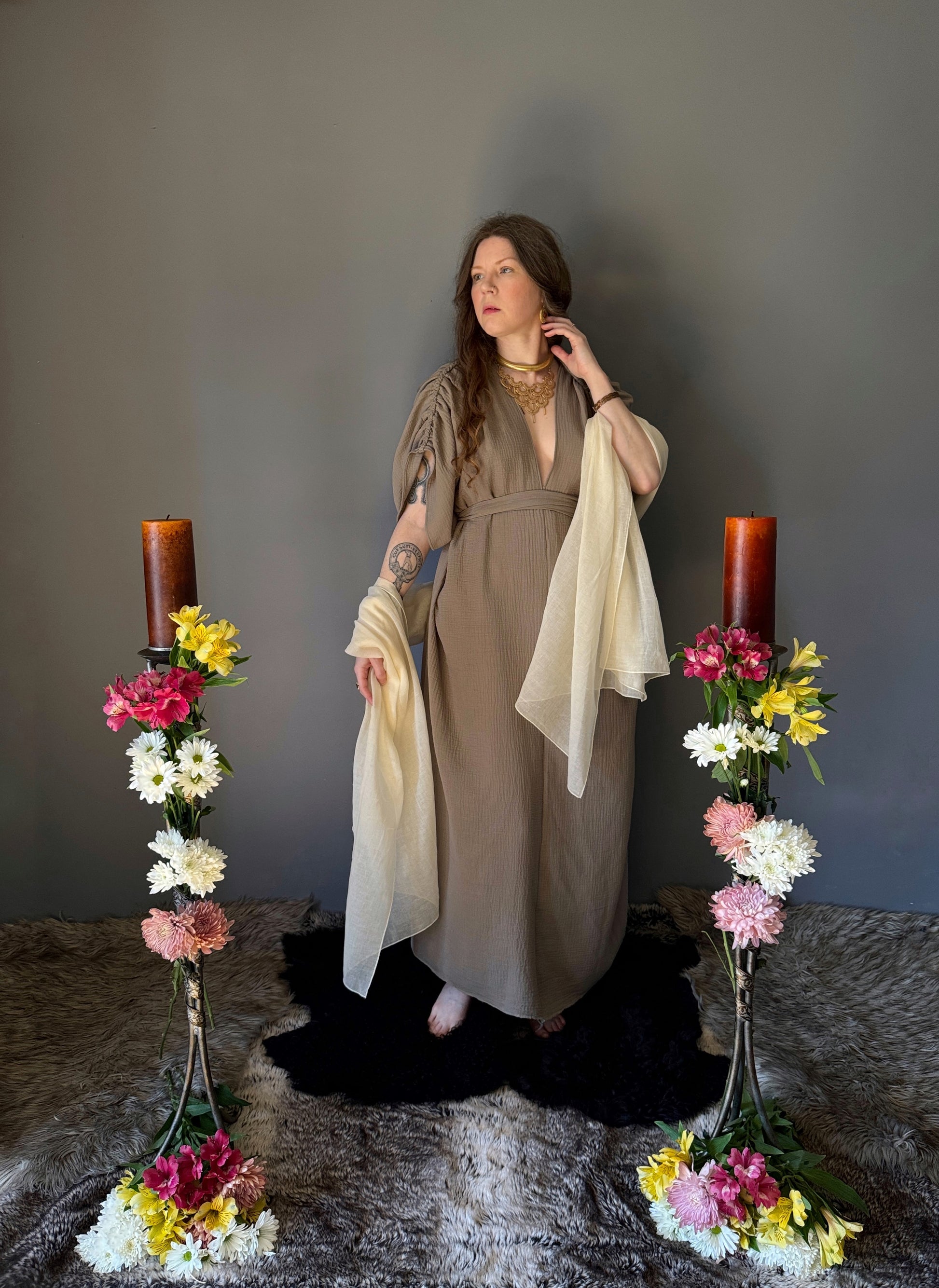 A priestess surrounded by flowers wearing a ancient greek inspired ritual robe. Featuring a deep v neck line, gathered short sleeves with braided trim that ties at the back. Cinched at the waist with a matching long wrap belt. The gown is a soft tan colour cotton with a slight waffle comb texture. The veil is made from a cotton gauze in a natural undyed state. For the magically inclined, witches, priestess, priest, pagan, hellenistic, polytheistic, spiritual or lover of history. Sustainably made in Canada