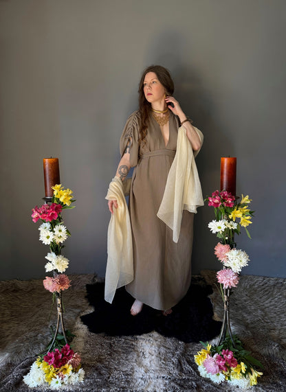 A priestess surrounded by flowers wearing a ancient greek inspired ritual robe. Featuring a deep v neck line, gathered short sleeves with braided trim that ties at the back. Cinched at the waist with a matching long wrap belt. The gown is a soft tan colour cotton with a slight waffle comb texture. The veil is made from a cotton gauze in a natural undyed state. For the magically inclined, witches, priestess, priest, pagan, hellenistic, polytheistic, spiritual or lover of history. Sustainably made in Canada