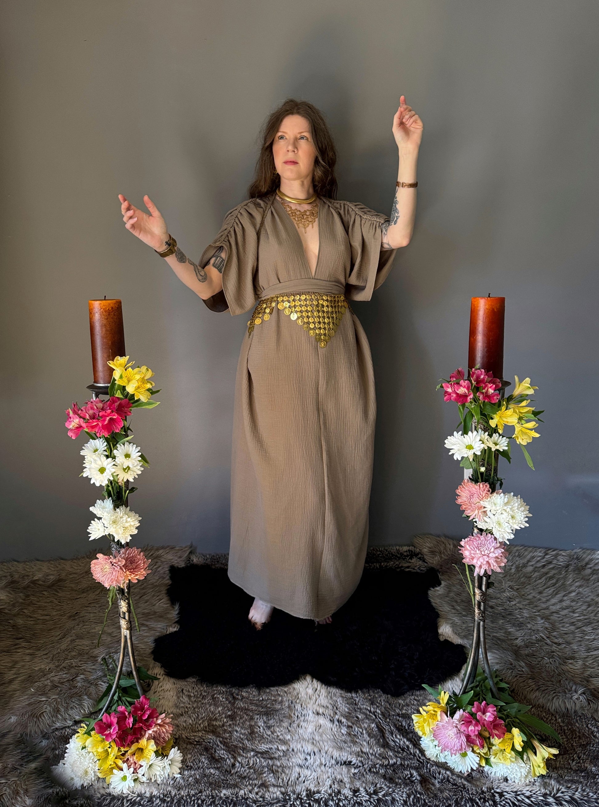 A priestess surrounded by flowers wearing a ancient greek inspired ritual robe. Featuring a deep v neck line, gathered short sleeves with braided trim that ties at the back. Cinched at the waist with a matching long wrap belt. The gown is a soft tan colour cotton with a slight waffle comb texture. The veil is made from a cotton gauze in a natural undyed state. For the magically inclined, witches, priestess, priest, pagan, hellenistic, polytheistic, spiritual or lover of history. Sustainably made in Canada