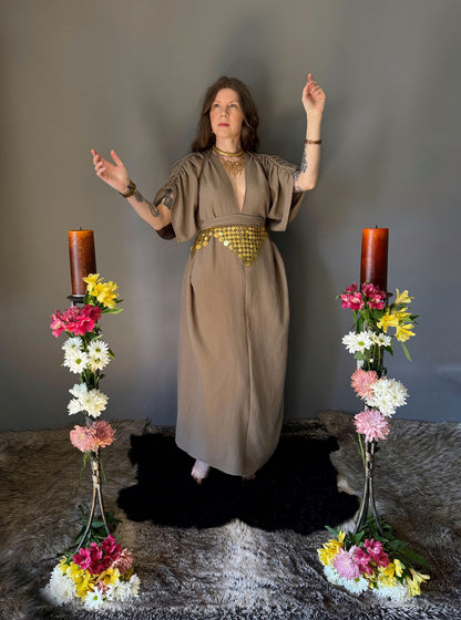 A priestess surrounded by flowers wearing a ancient greek inspired ritual robe. Featuring a deep v neck line, gathered short sleeves with braided trim that ties at the back. Cinched at the waist with a matching long wrap belt. The gown is a soft tan colour cotton with a slight waffle comb texture. The veil is made from a cotton gauze in a natural undyed state. For the magically inclined, witches, priestess, priest, pagan, hellenistic, polytheistic, spiritual or lover of history. Sustainably made in Canada
